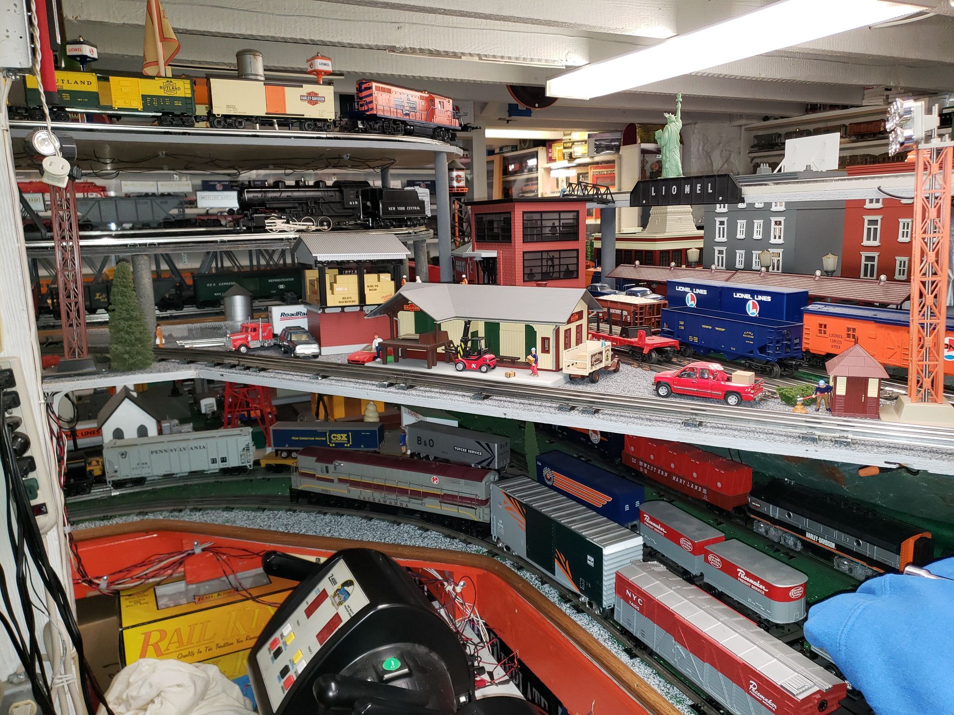 Indoor model train layout in a cluttered workshop with red and green tracks and miniature buildings.