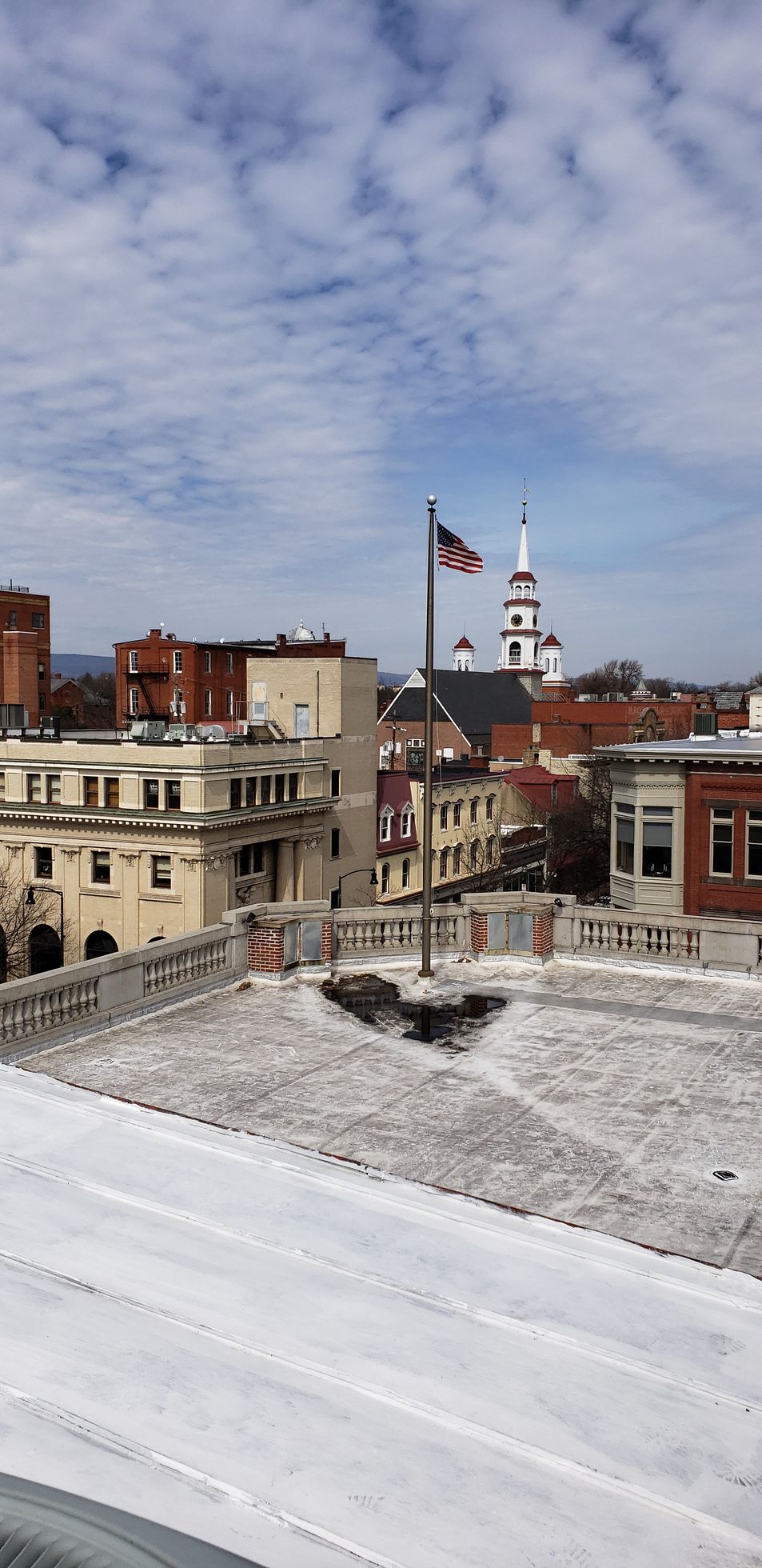 Snowy rooftop view of a city skyline with brick buildings, a flagpole, and church steeples under a cloudy sky