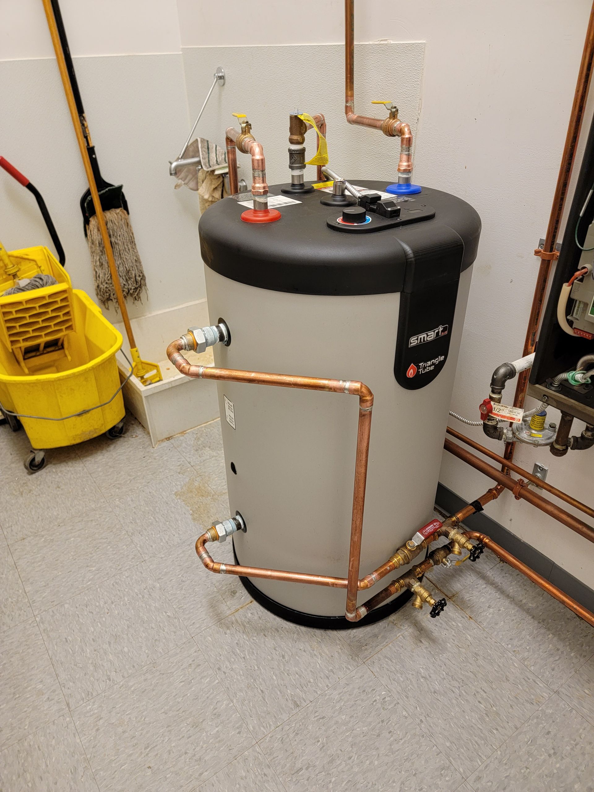 Gray water heater with copper pipes and valves in a utility room, beside a yellow bucket and mop.