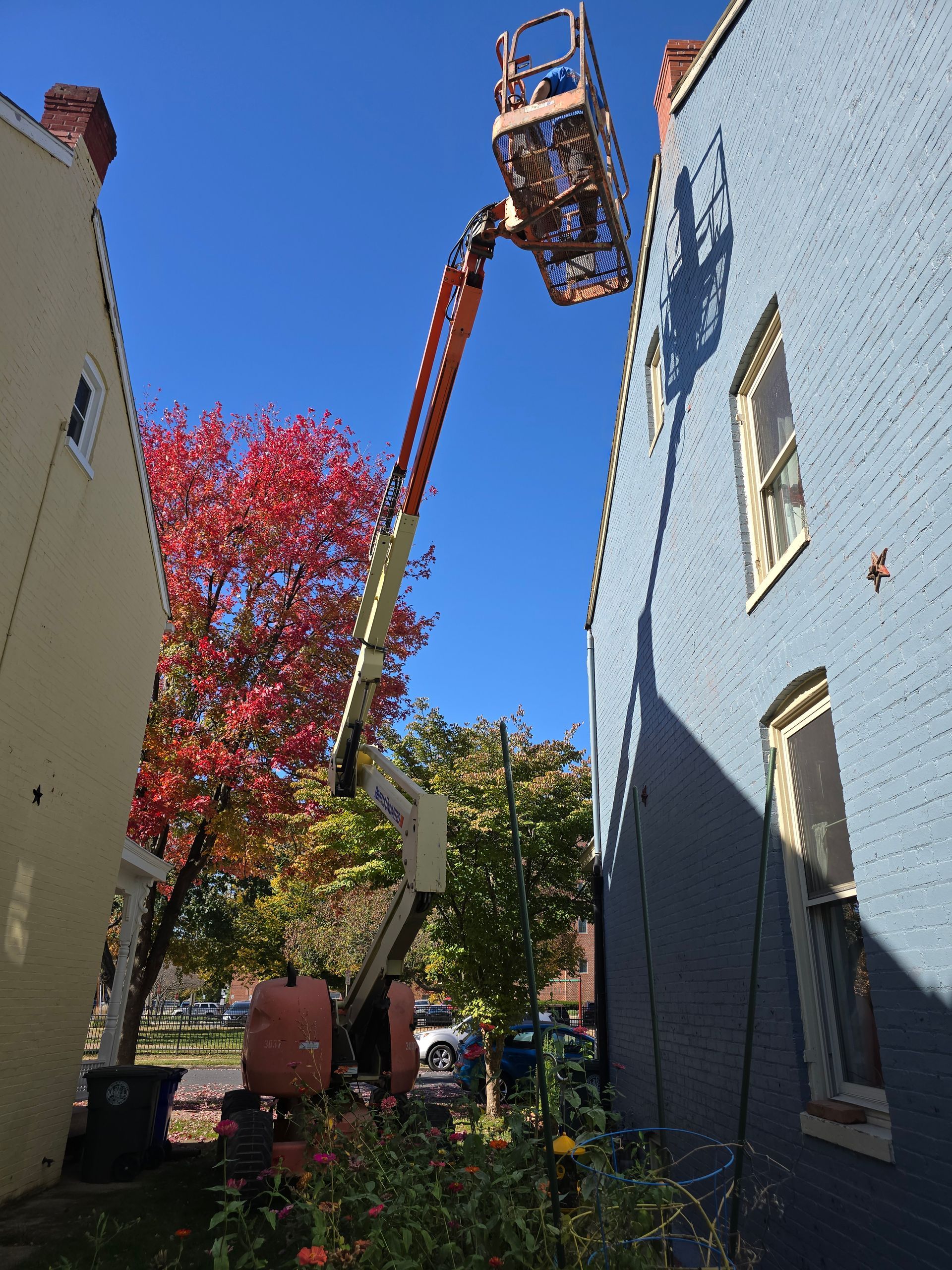 Crane reaching between buildings over a garden, with blue sky and autumn trees.