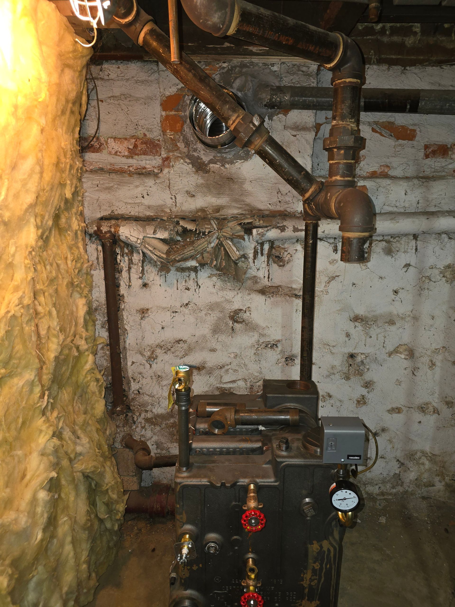 Old boiler and exposed pipes in a basement against a rough brick wall.