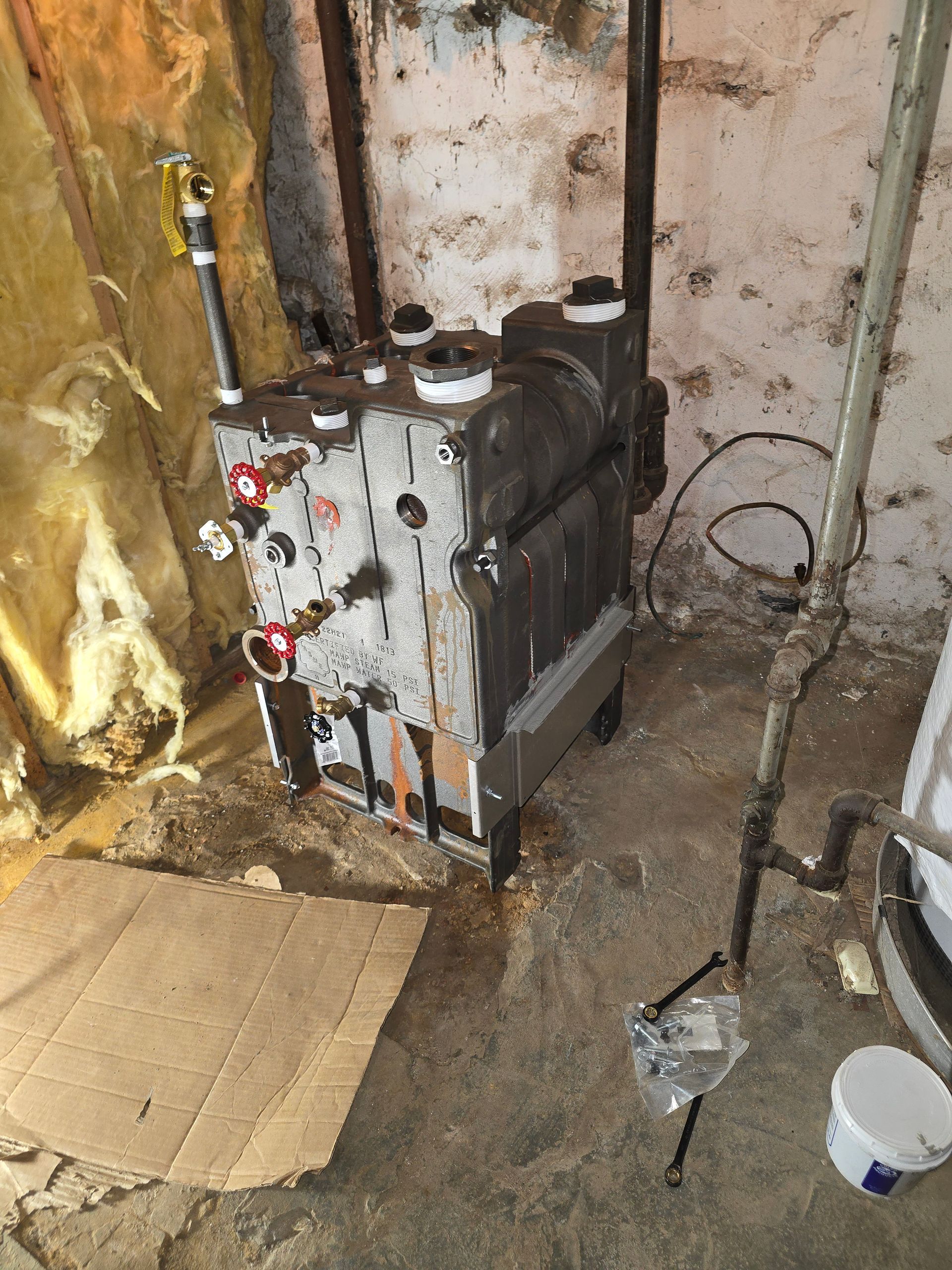 Basement utility area with a rusty water heater, pipes, and a bucket on the floor.