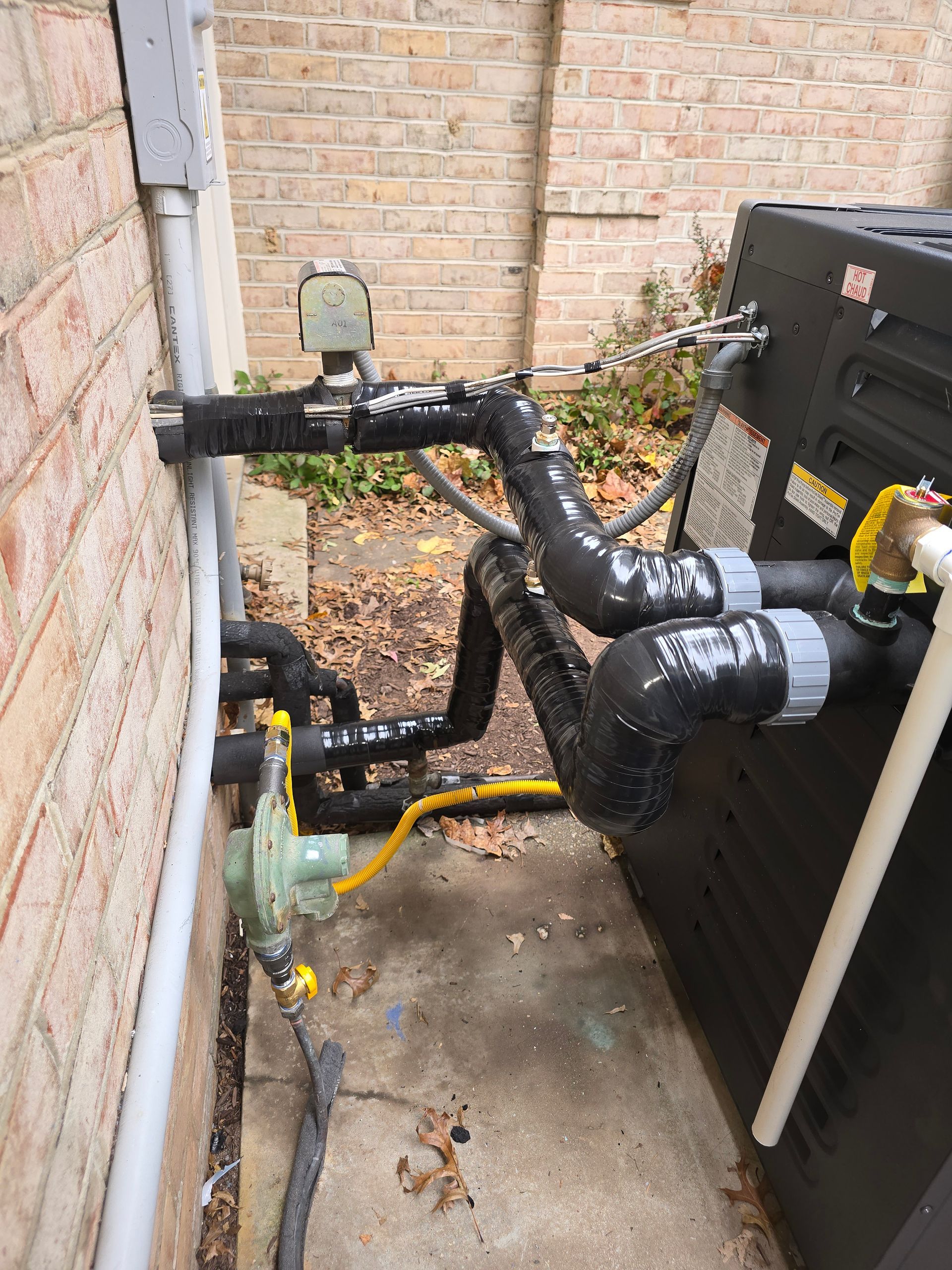 Outdoor HVAC equipment with black insulated pipes and valves beside a brick wall and fence.