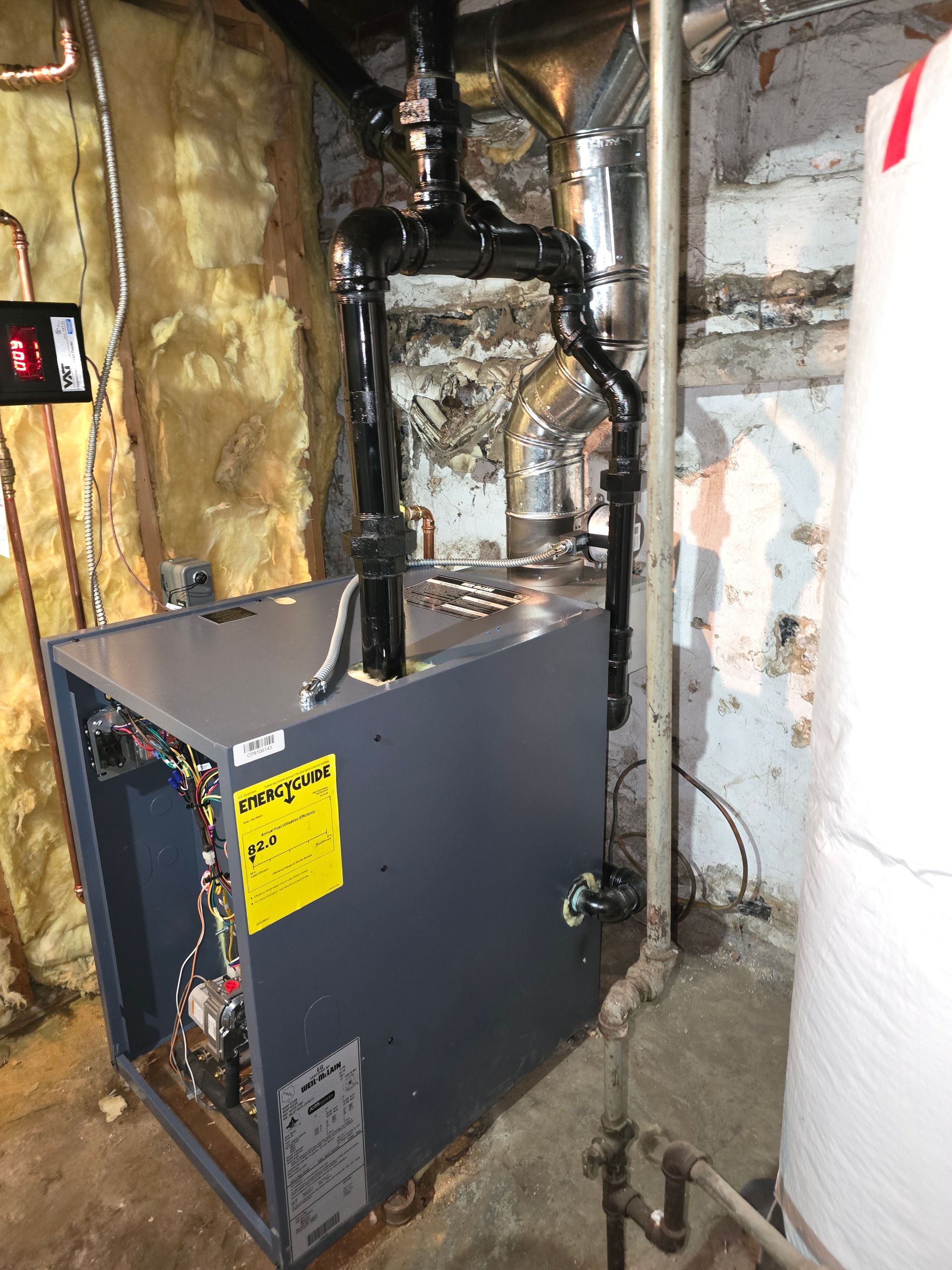 Gray furnace in a basement with exposed pipes, insulation, and unfinished stone walls.