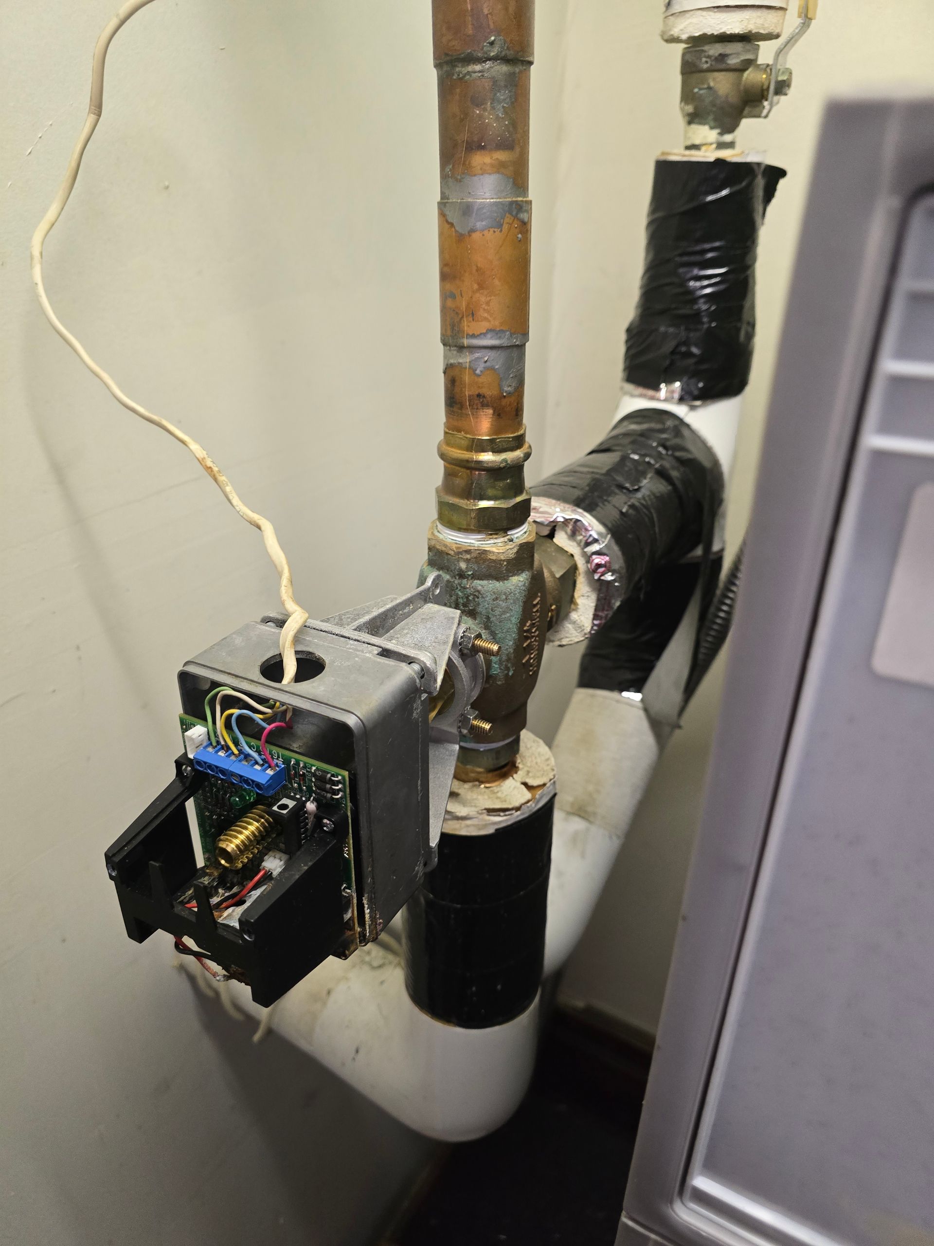 Open electrical control box mounted on indoor plumbing pipes with taped black insulation and wire connected.