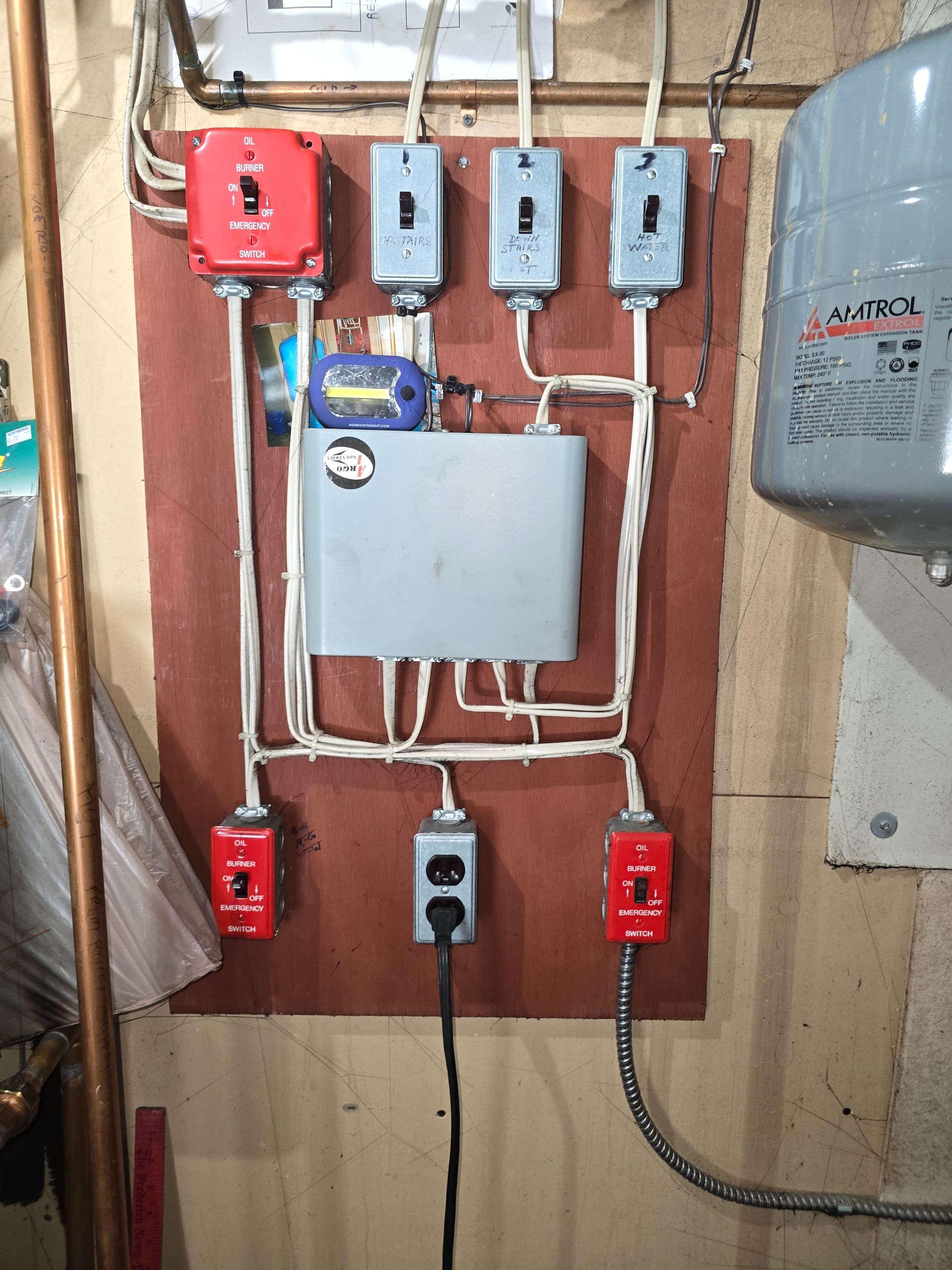 Electrical panel with switches and wiring mounted on a wooden board beside a gray tank and conduit pipes.