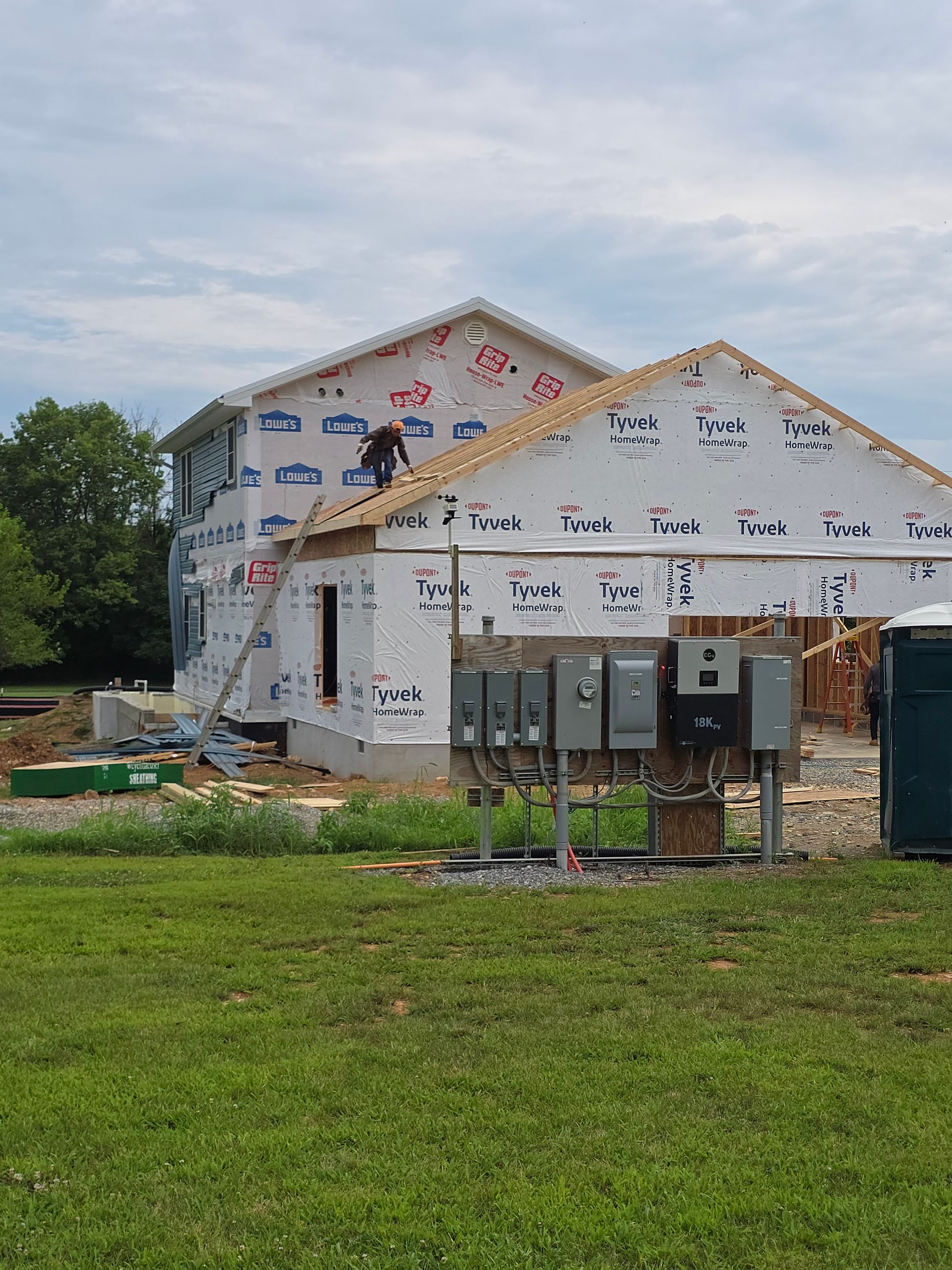 Partially built house wrapped in white housewrap with exposed framing and utility boxes in a grassy yard