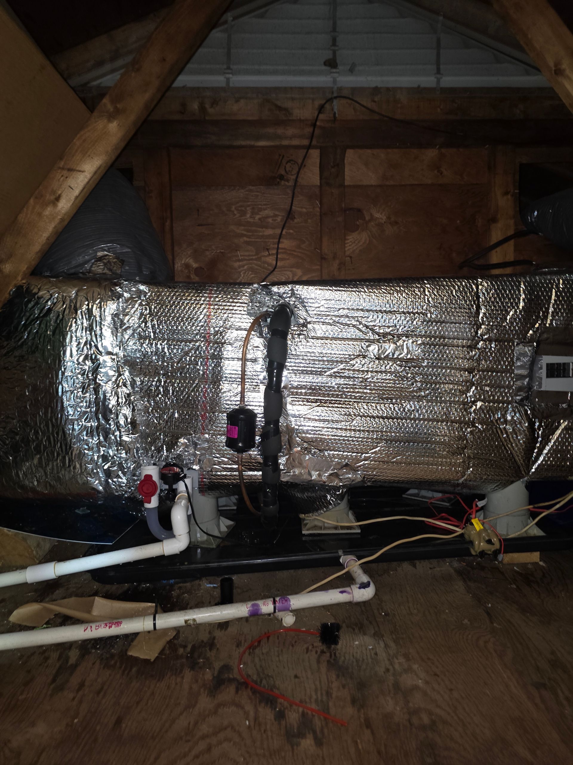 Attic crawlspace with foil-wrapped ductwork, exposed beams, and plumbing pipes under the roof.