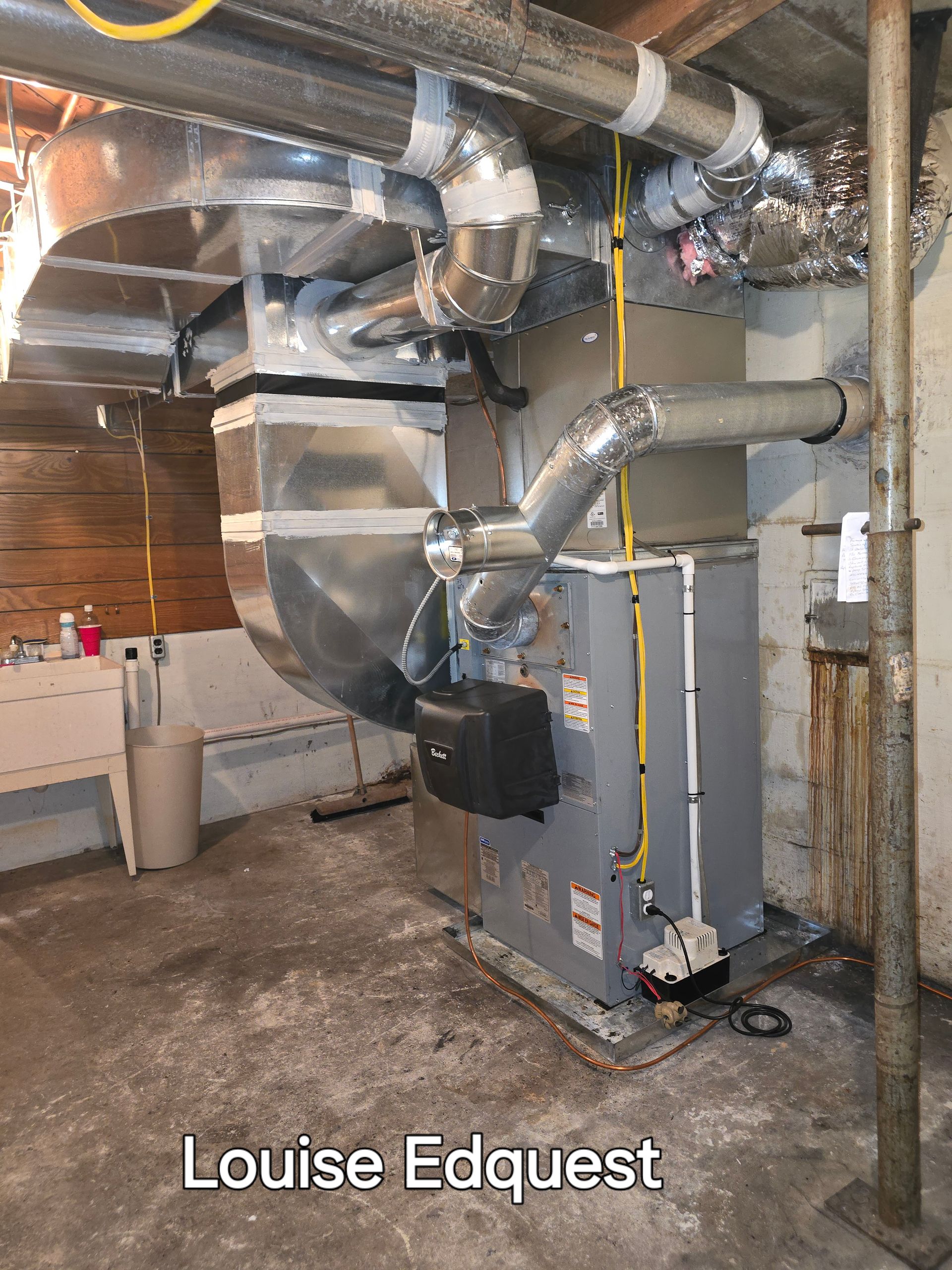 Basement utility room with large metal HVAC ducts and a gray furnace unit against exposed brick walls.