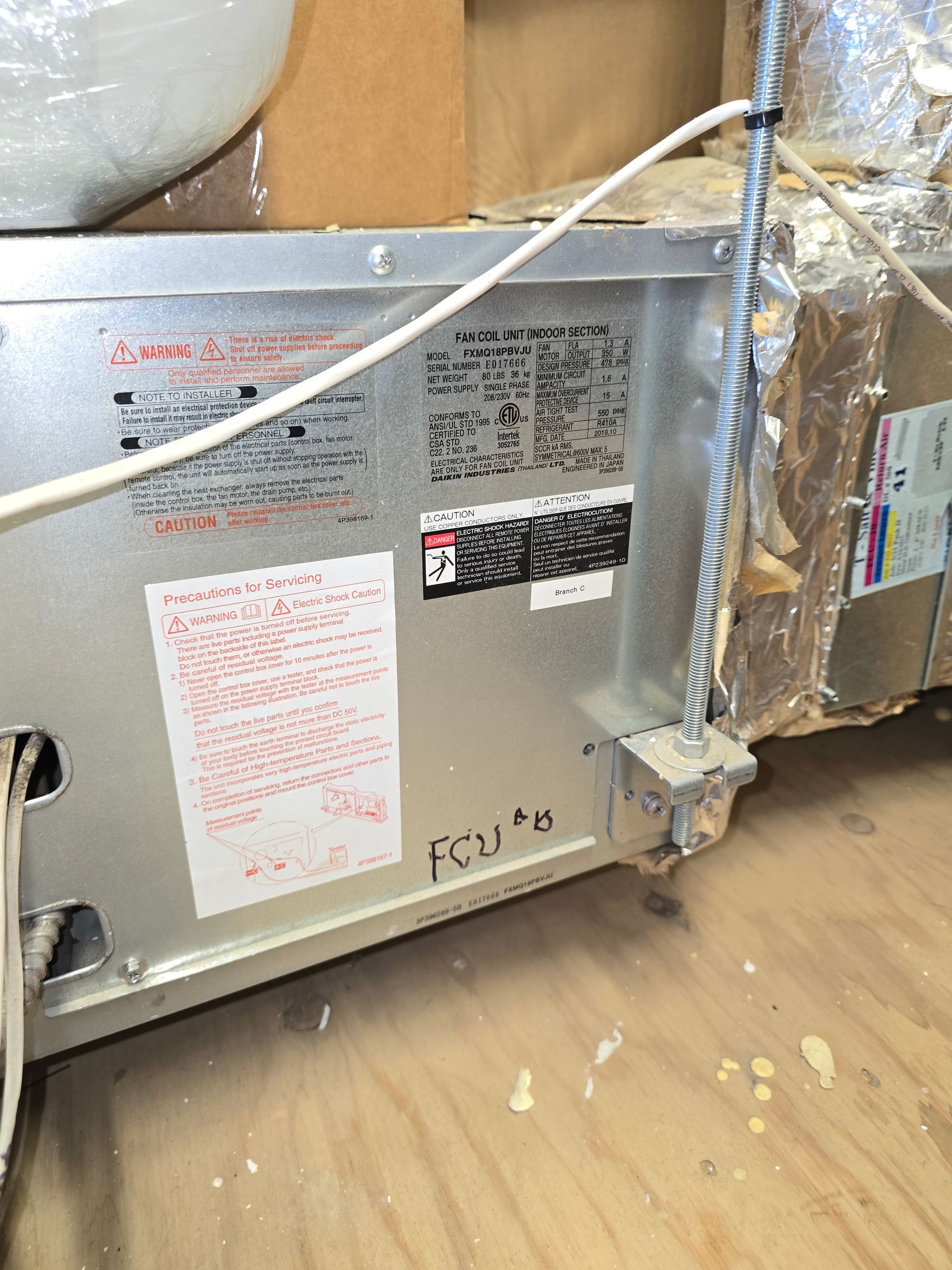 Metal HVAC unit with labels and a white cable, partially wrapped in plastic, in a storage area
