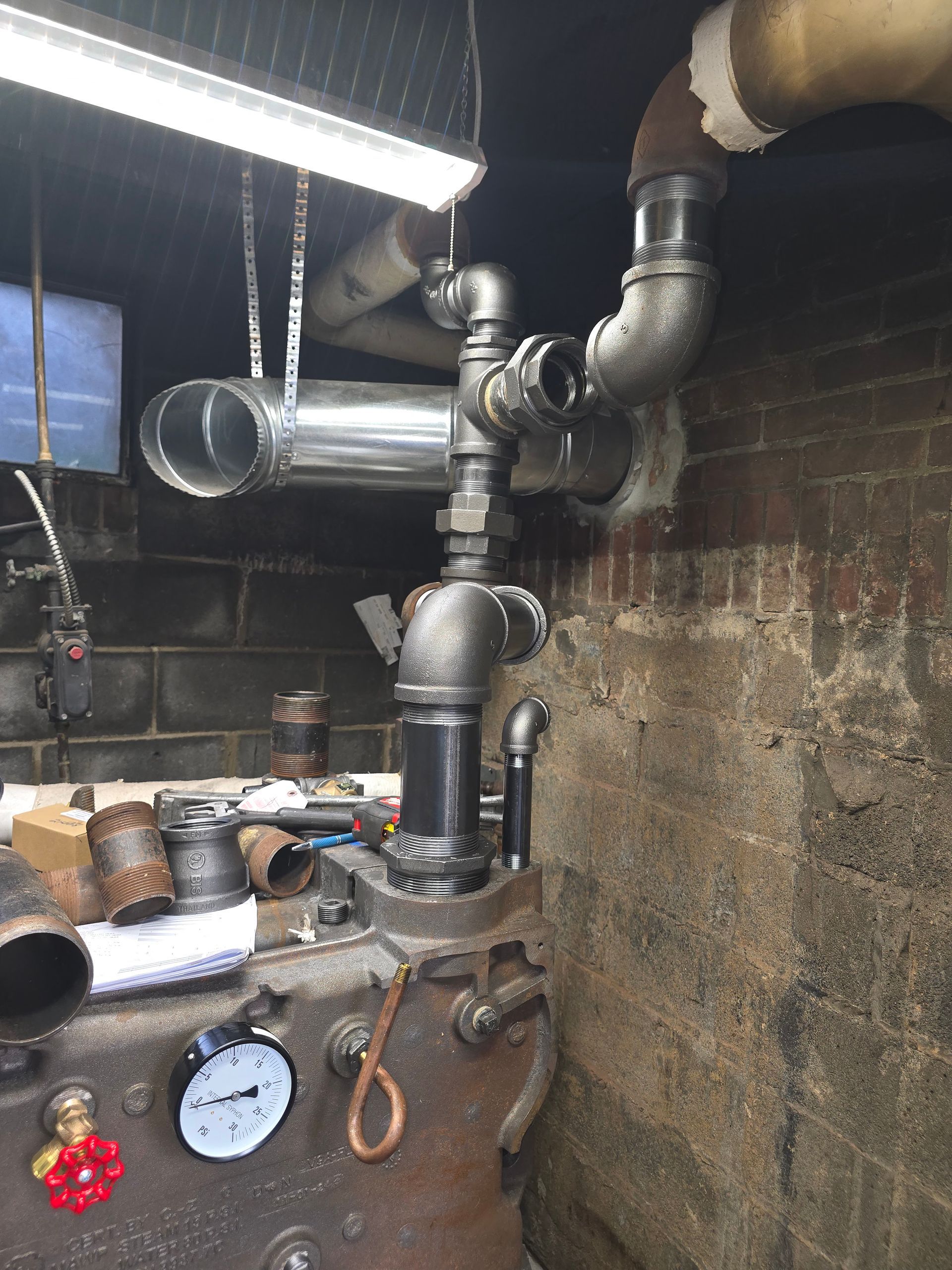 Industrial boiler room with metal pipes, valves, pressure gauge, and a brick wall under fluorescent lights