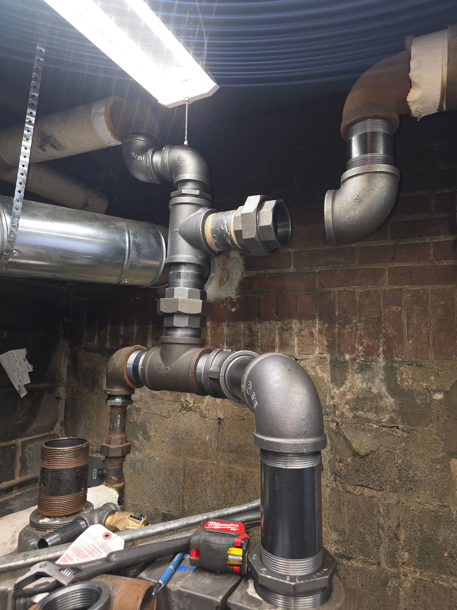 Basement plumbing with large metal ducts and pipes against a brick wall, under a bright fluorescent light.