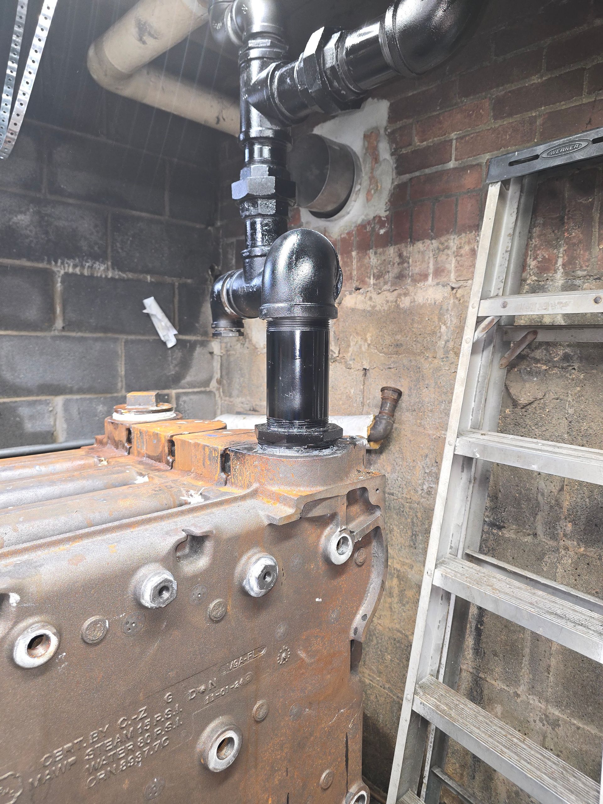 Rusty industrial equipment with black pipes in a brick utility room, ladder on the right
