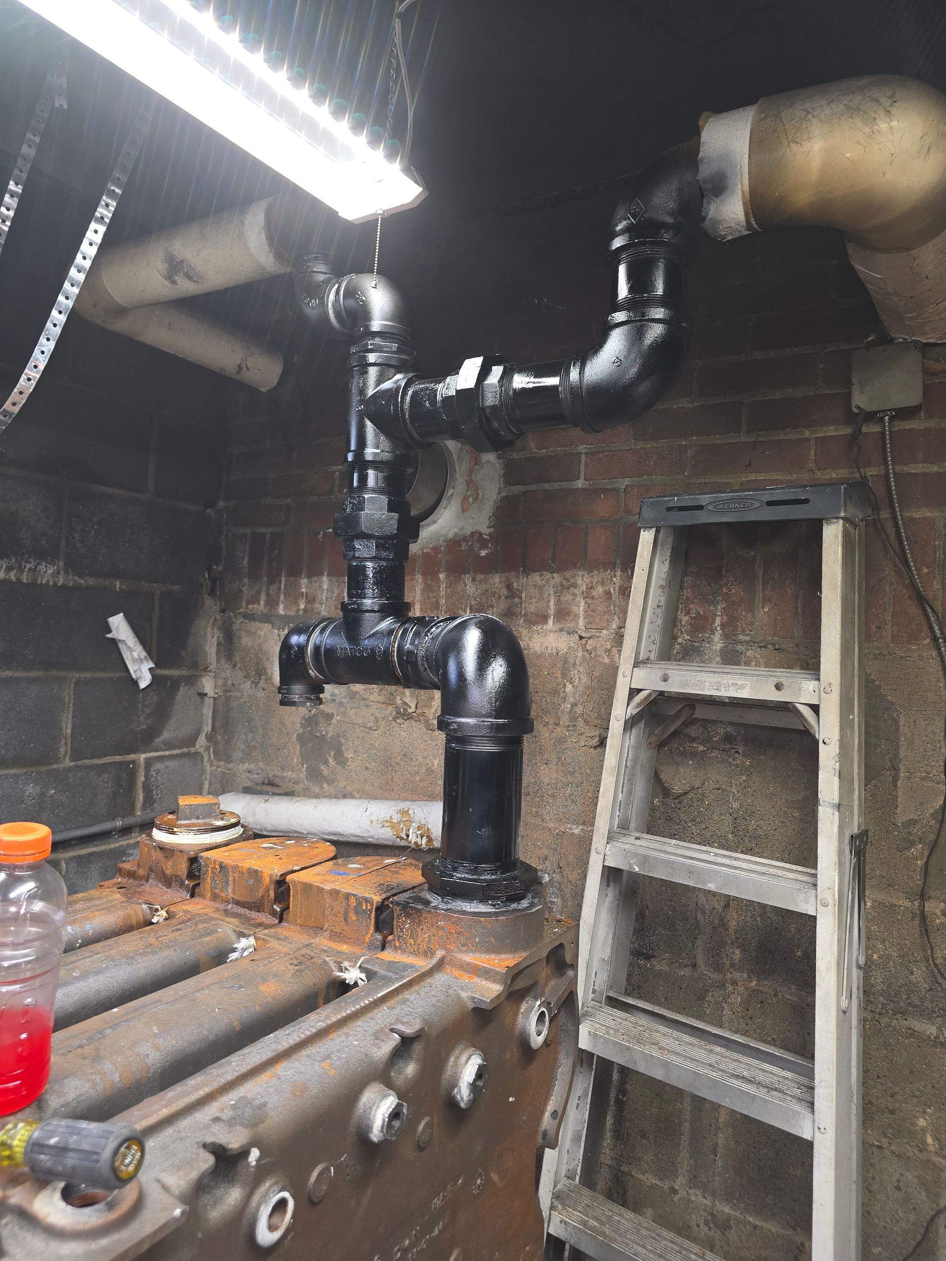 Basement utility room with black pipes, brick walls, a ladder, and a workbench under a fluorescent light