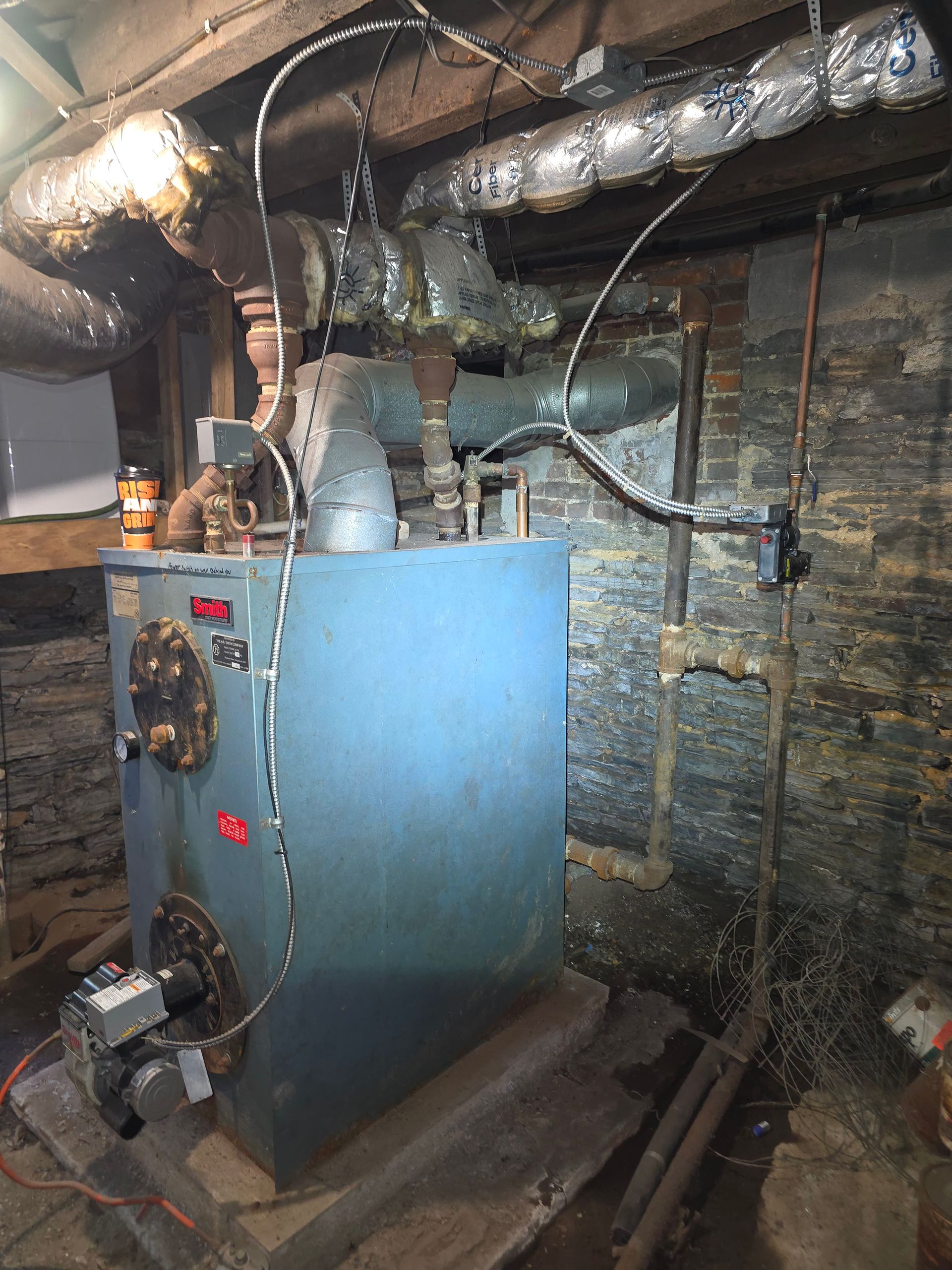 Blue boiler in a basement with exposed pipes and ductwork against brick walls