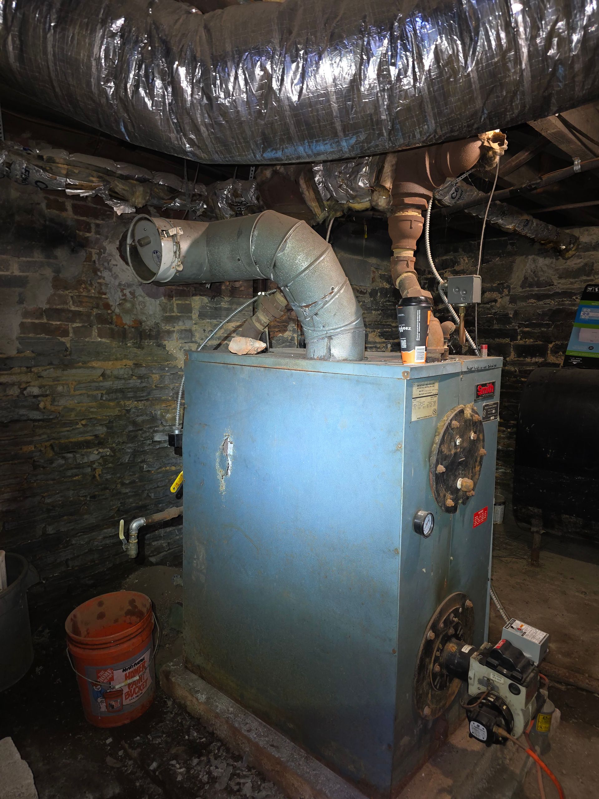 Blue boiler in a basement with large silver ducting and copper pipes overhead, near a red bucket.