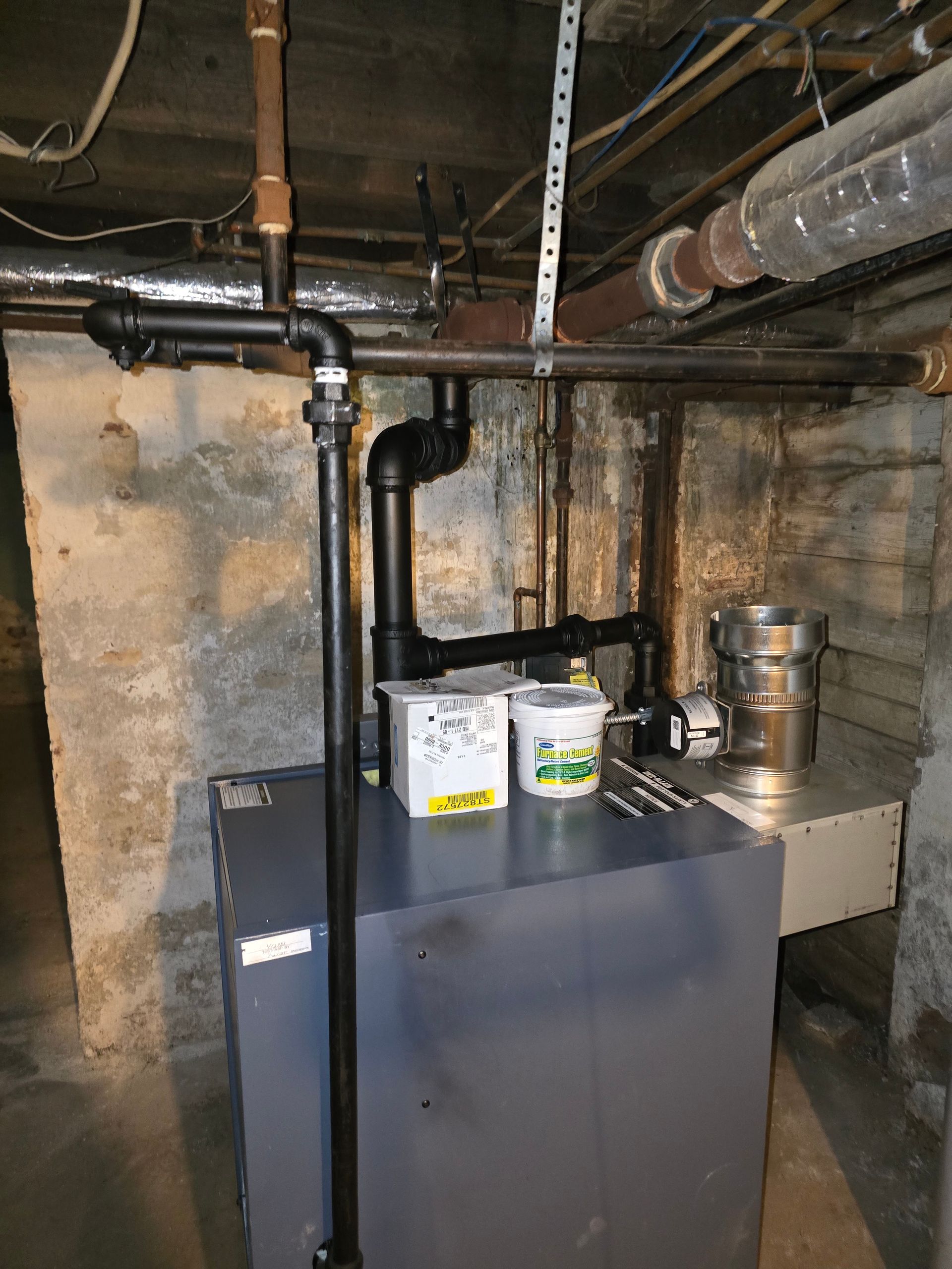 Basement utility room with gray boiler, black piping, exposed pipes, and paint cans on top.