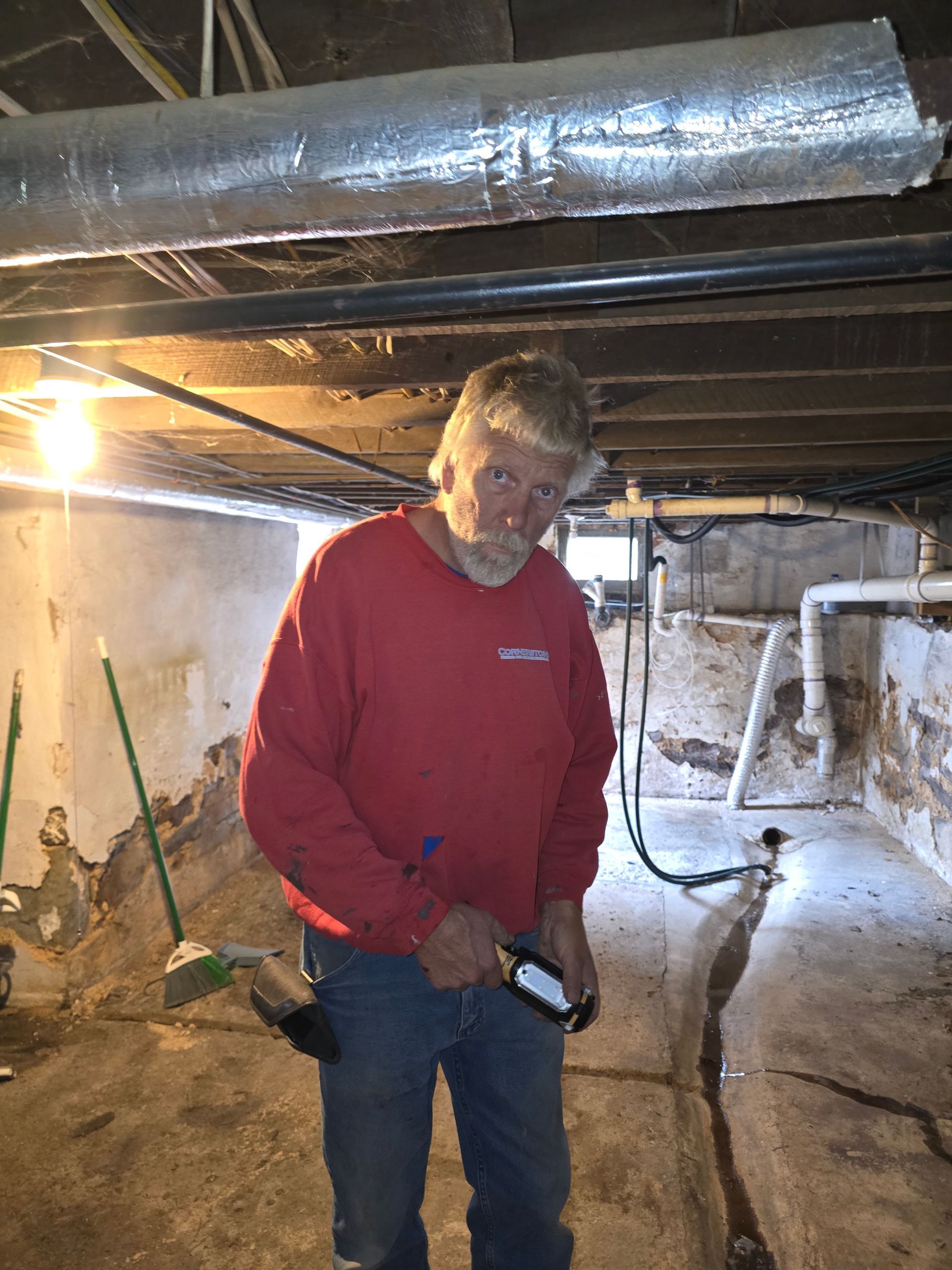 Person in a red sweatshirt using a flashlight in a dim crawlspace with pipes overhead.