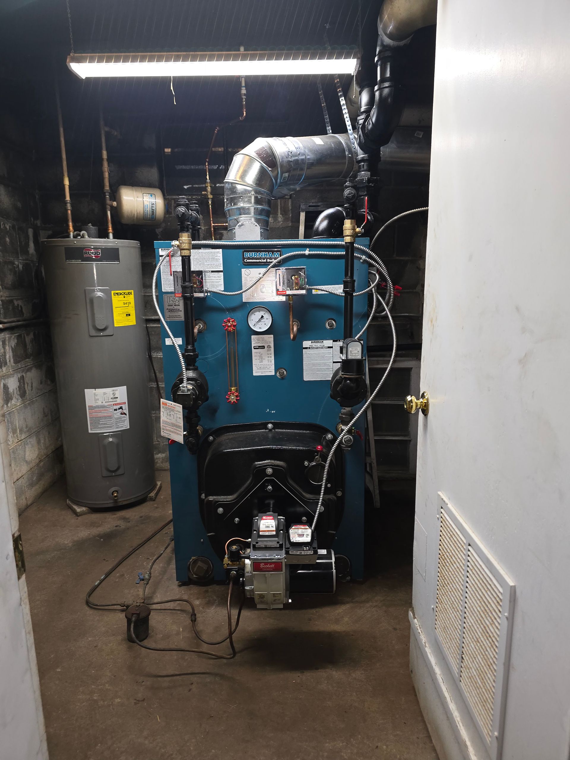 Commercial boiler room with a large blue boiler, water tank, and metal ductwork in a narrow utility space