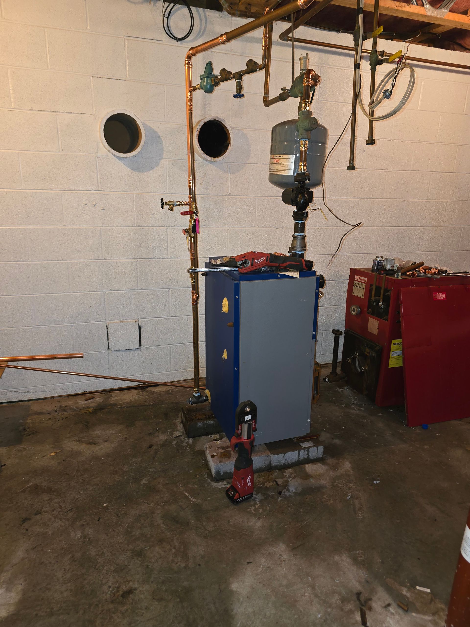 Basement utility room with blue and red boilers, copper piping, and concrete walls.