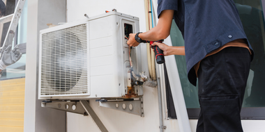 Technician repairing an outdoor air conditioner unit with tools on a building wall