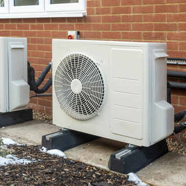 Outdoor air conditioning unit beside a brick house, mounted on black pads with visible pipes and vents
