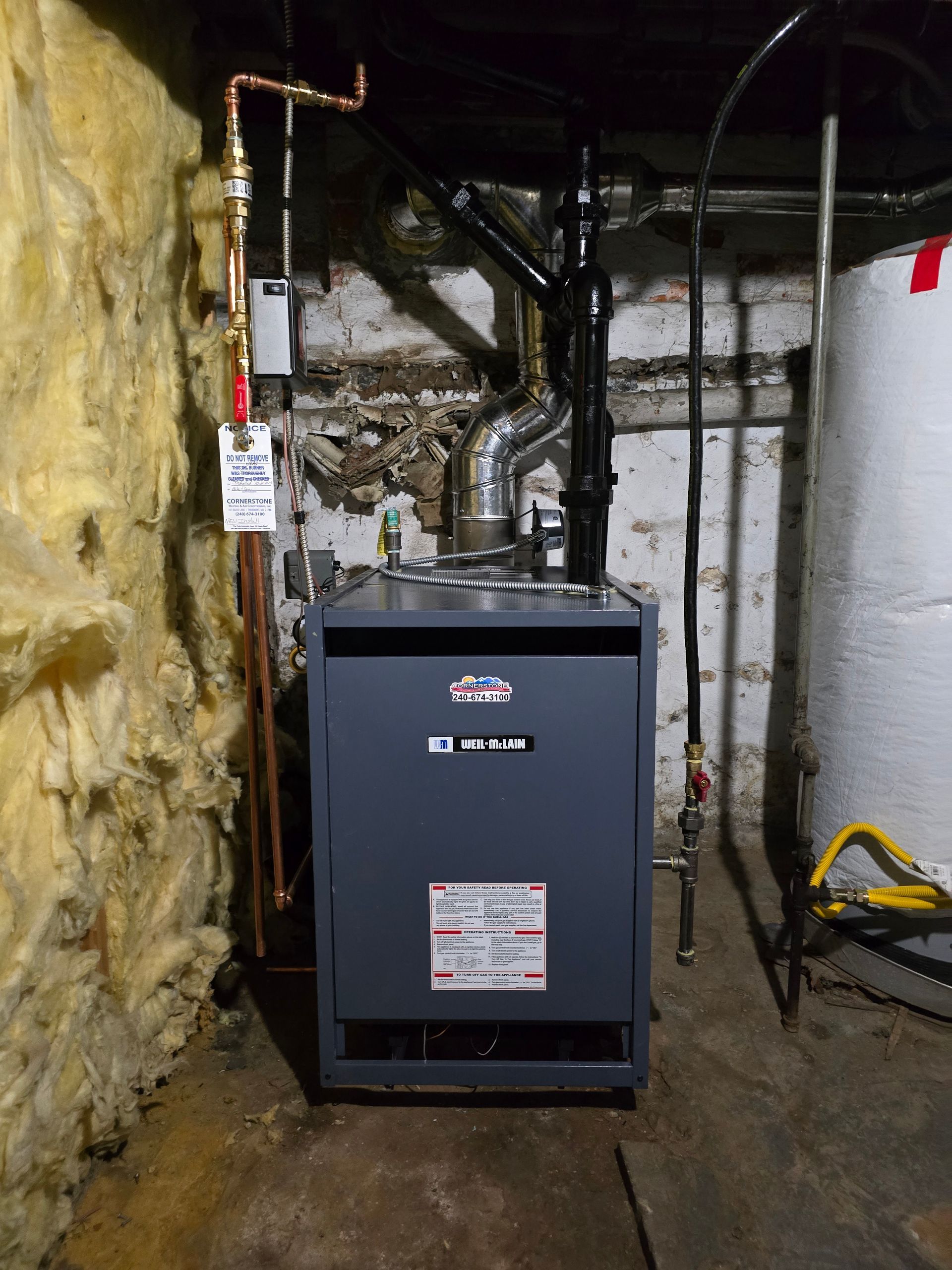 Gray furnace in a narrow basement utility space with exposed insulation and pipes.