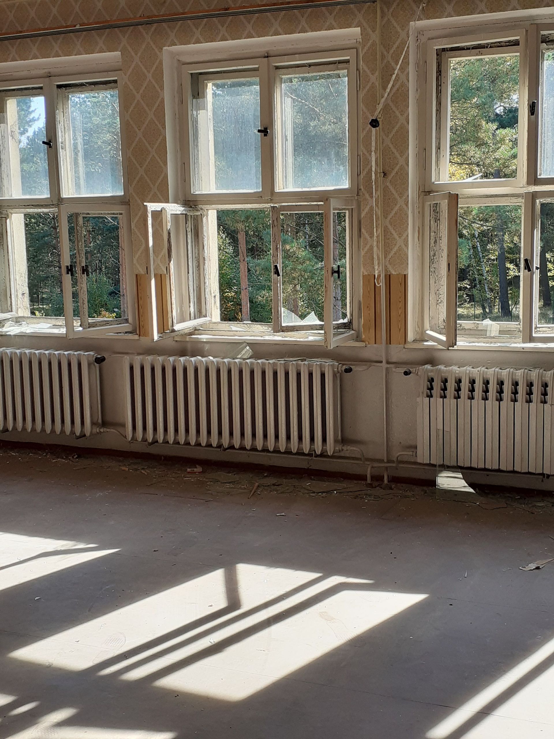 Sunlit empty room with large windows, radiators, and long shadows on the floor.