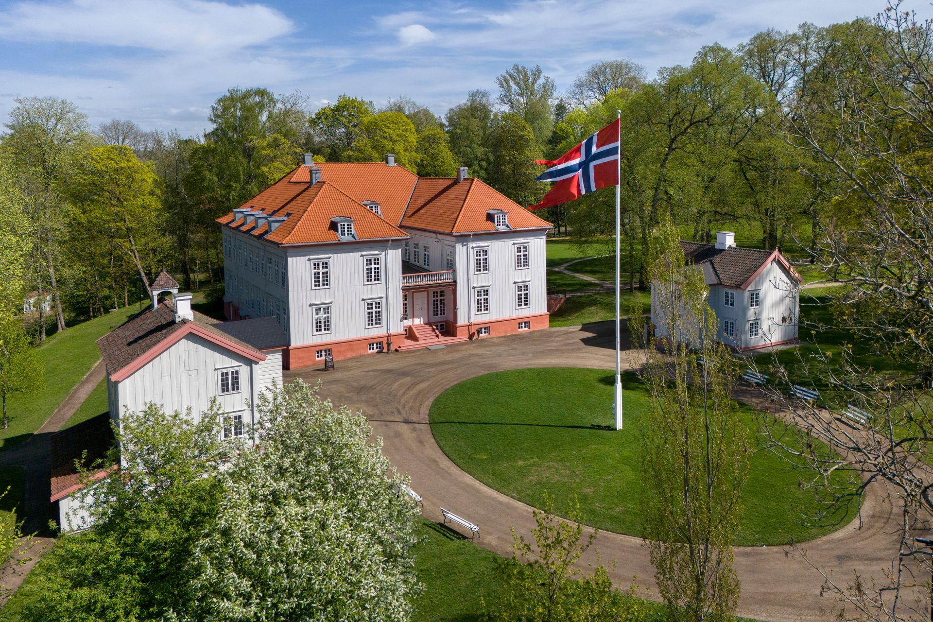 Hvitt herregård med rødt tak og norsk flagg på en sirkulær innkjørsel, omgitt av trær og grøntområder.