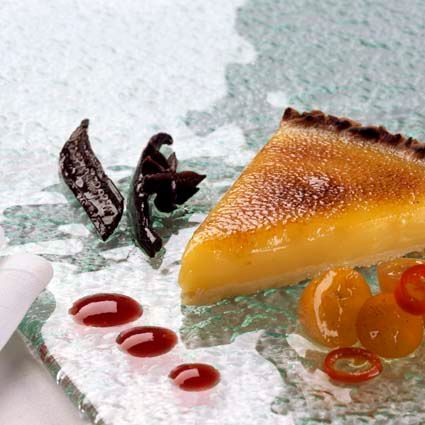 En skive sitronterte på en teksturert glassplate, garnert med stjerneanis, vaniljestang, røde prikker med saus og frukt.
