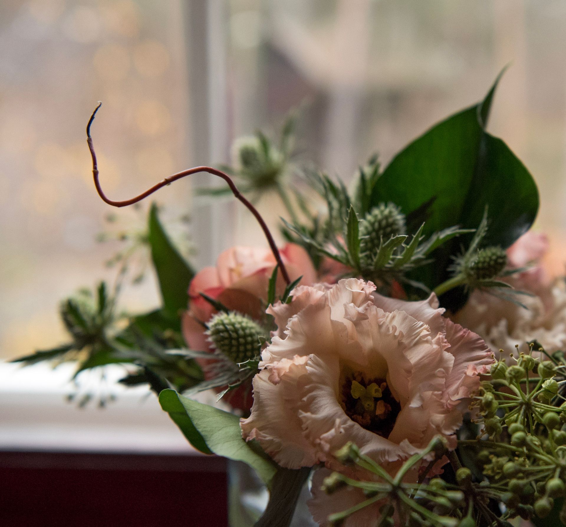 Et nærbilde av et blomsterarrangement med rufsete, bleke ferskenroser, piggete blågrønne tistelblomster og tynne, krøllete ranker.