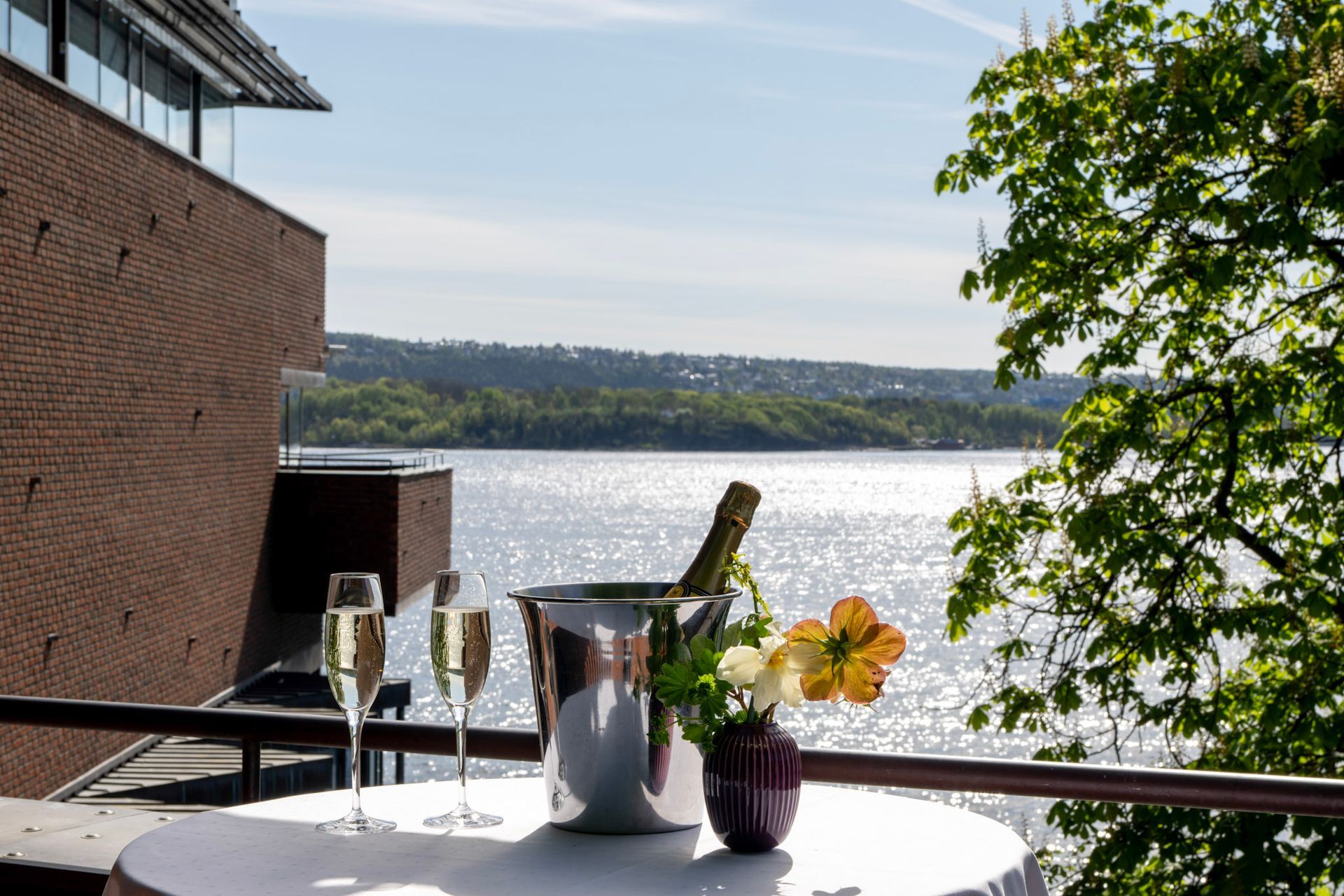 To champagnefløyter, en isbøtte med en flaske og blomster på et bord med utsikt over en innsjø ved en murbygning.