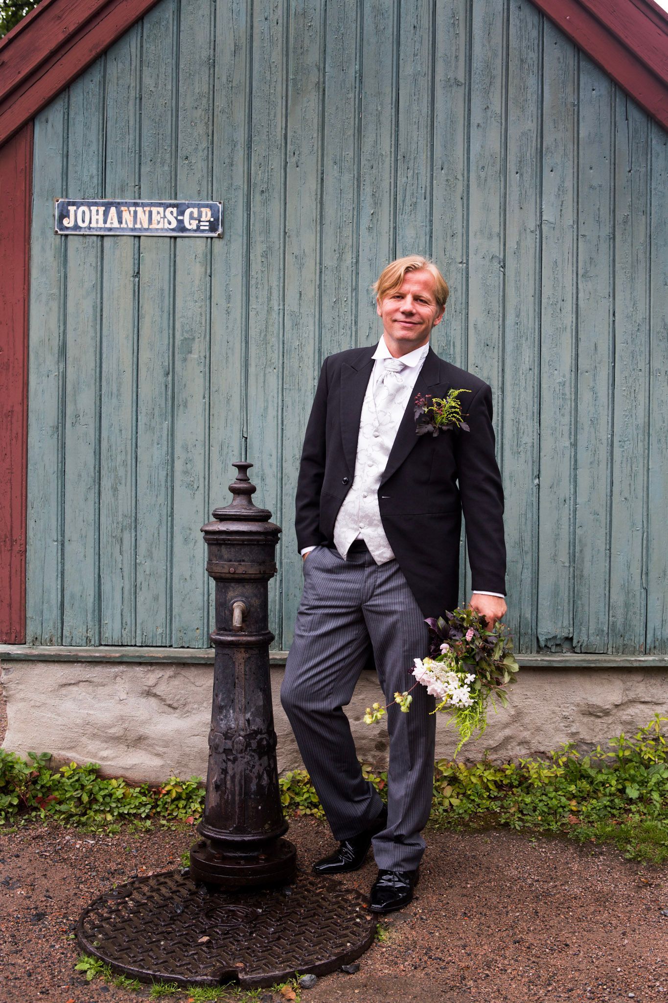 Mann i mørk dress lener seg ved siden av en svart stolpe foran en forvitret grønn låvedør, med blomster