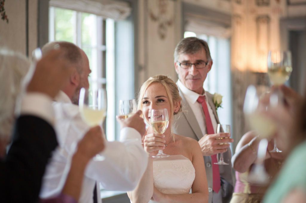 Folk på en bryllupsfest skåler med champagne i et sterkt opplyst, dekorert rom.