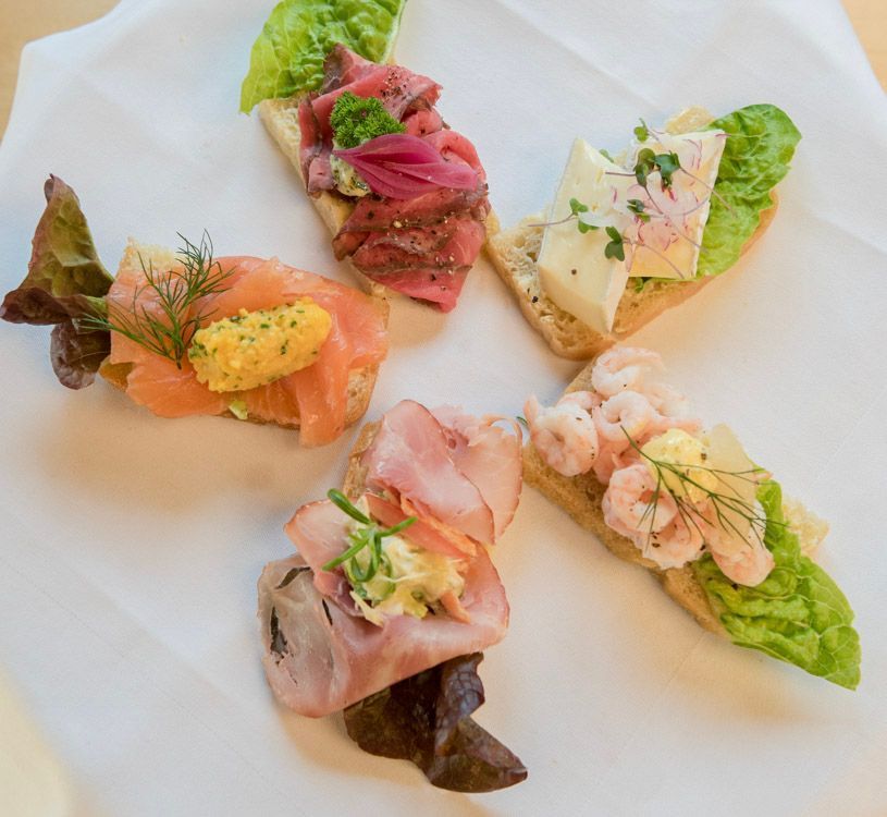 Fem åpne smørbrød med variert topping som laks, roastbiff, reker, skinke og ost arrangert på en tallerken.