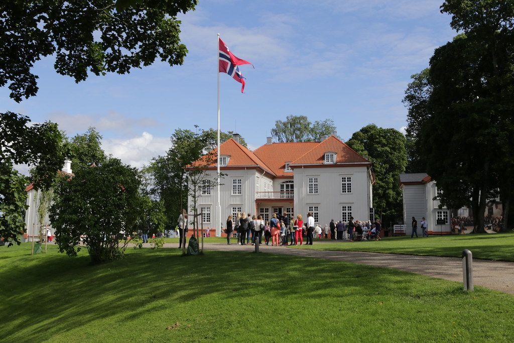 Stor hvit bygning med norsk flagg, folk samlet på plen, trær, solskinnsdag.