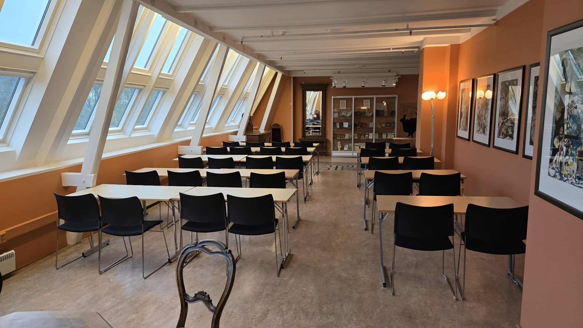 Et tomt, sterkt opplyst møterom med rader med bord og svarte stoler, skråstilte vinduer og kunst på oransje vegger.