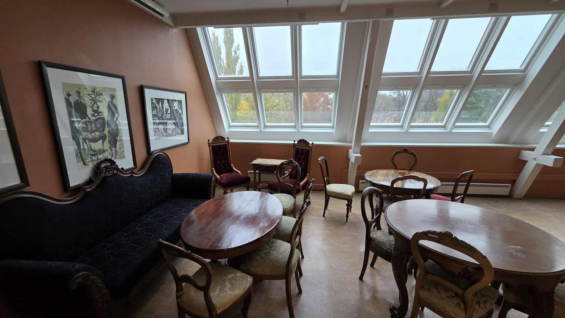 Et koselig, rustikt loftsrom med skråstilte takvinduer, antikke møbler, mørke trebord og innrammet kunst på oransje vegger.