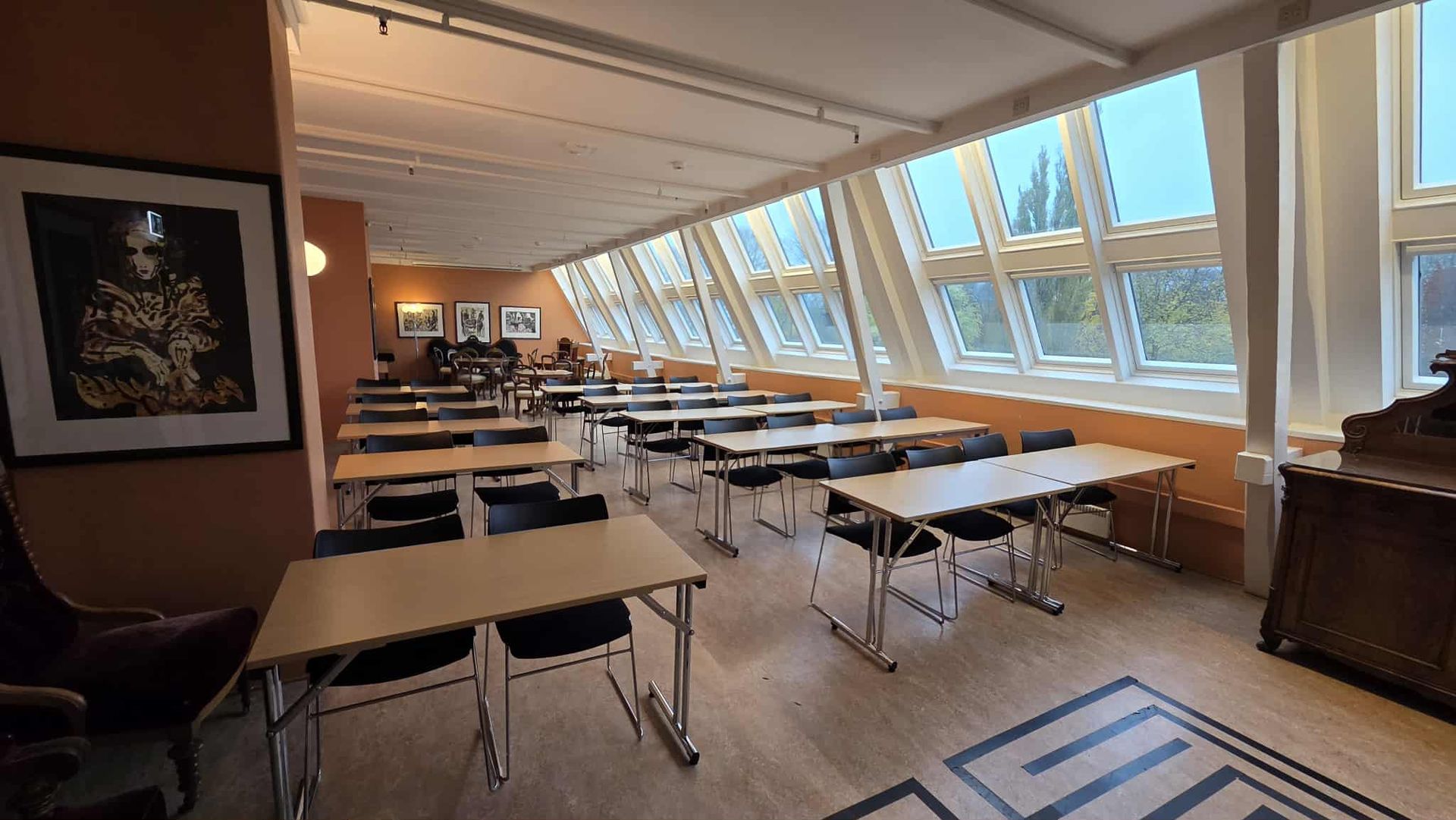 Et lyst, moderne opplæringsrom med rader med bord og stoler, store skråstilte vinduer og et innrammet trykk på veggen.