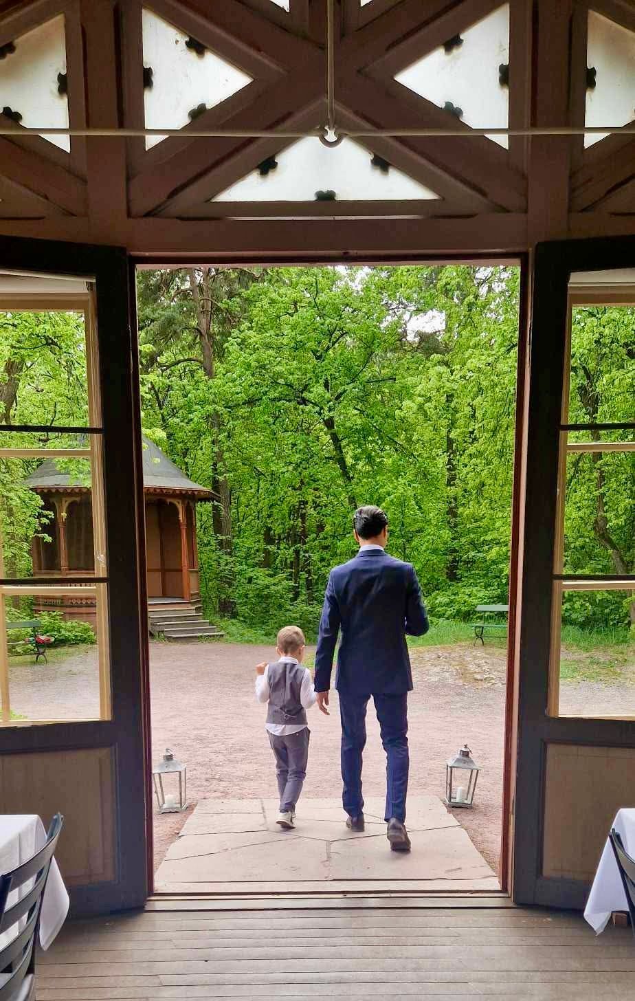 En person i dress og et barn går gjennom en åpen døråpning mot en frodig, grønn park.