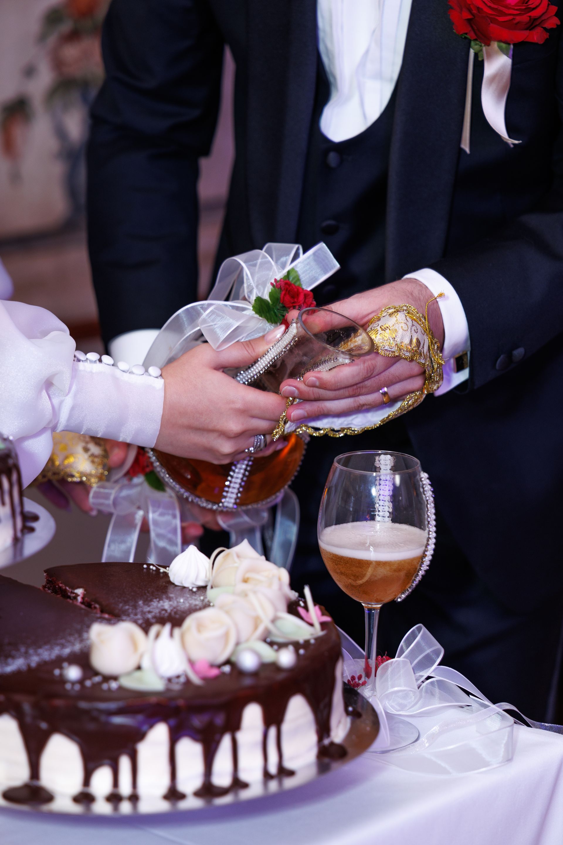 Et brud og brudgom heller en drink i et glass ved siden av en bryllupskake med sjokoladeglasur.