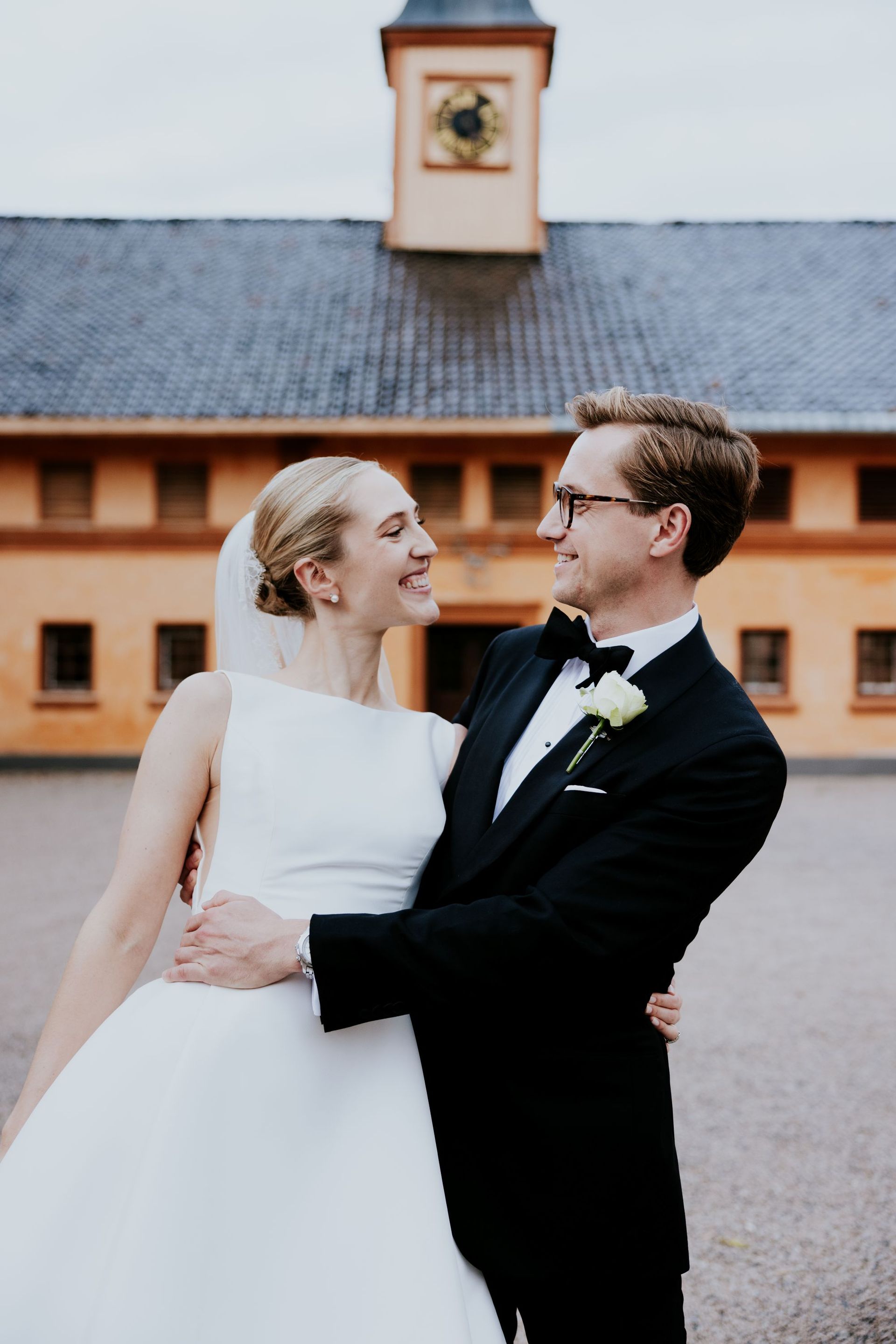En brud i hvit brudekjole og en brudgom i svart smoking smiler til hverandre utenfor en bygning med et klokketårn.