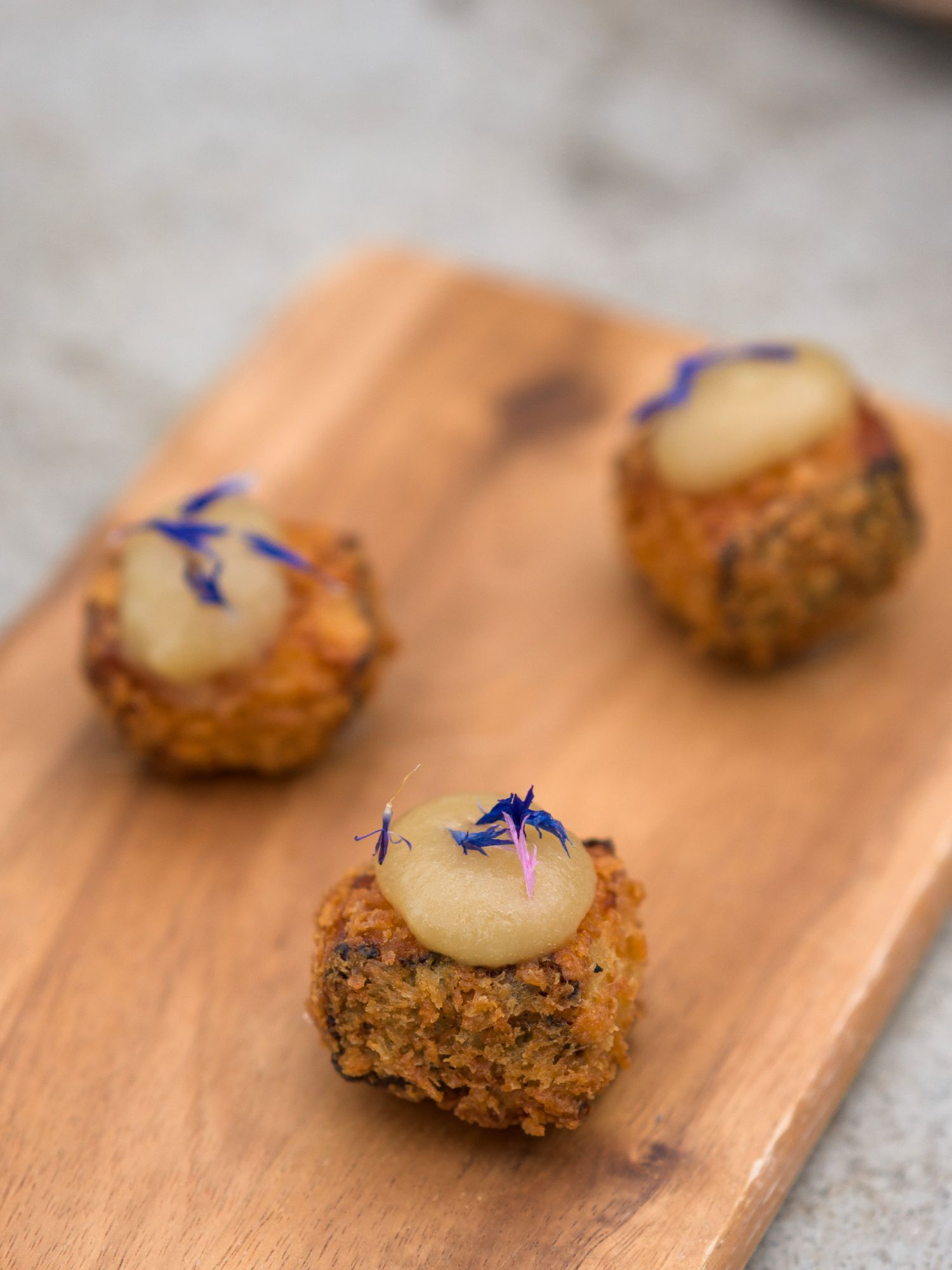 Tre gyllenbrune forretter toppet med klatter av lett saus og blå spiselige blomster, arrangert på en treplate.