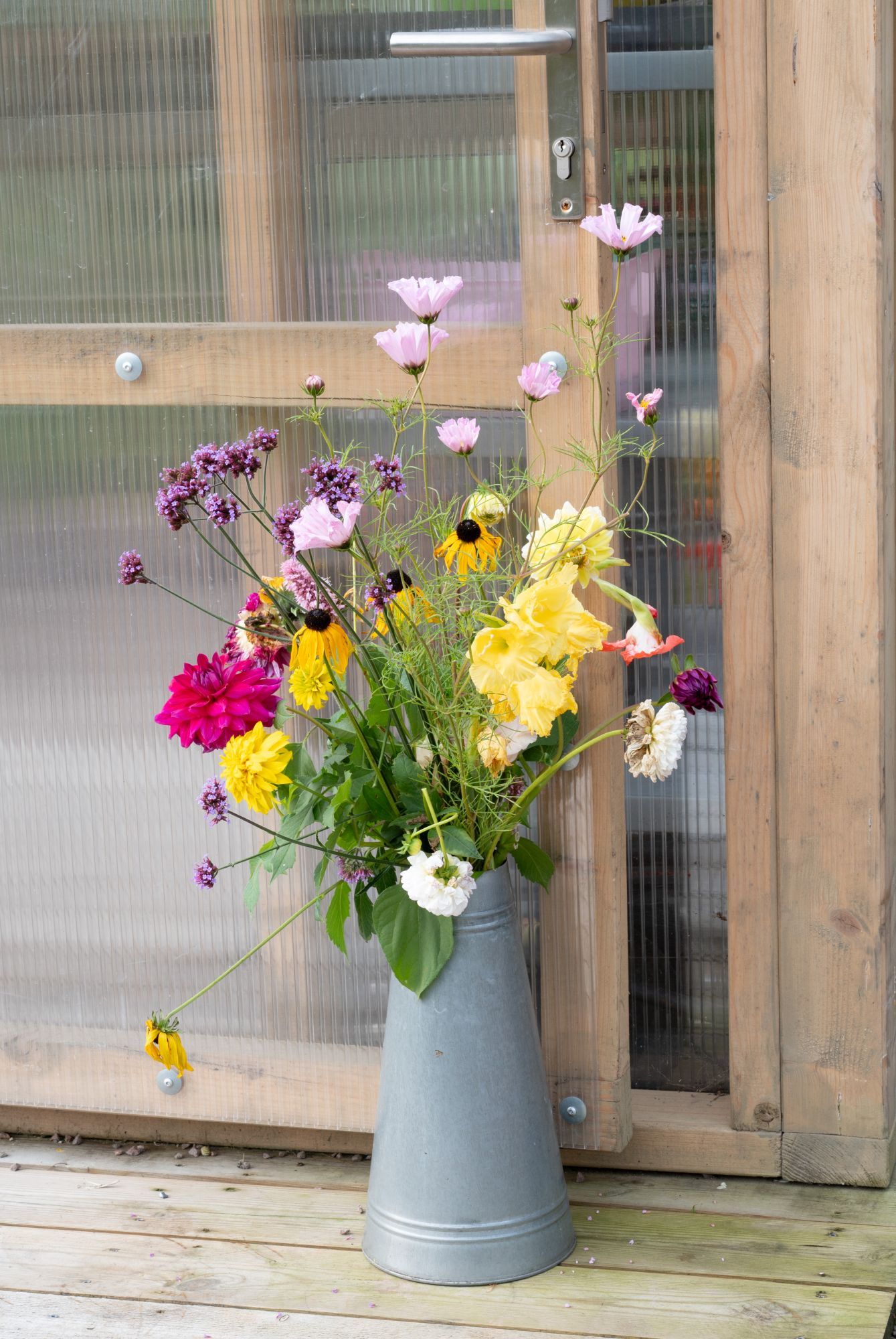 Bukett med fargerike markblomster i en høy grå vase på en treveranda ved en dør