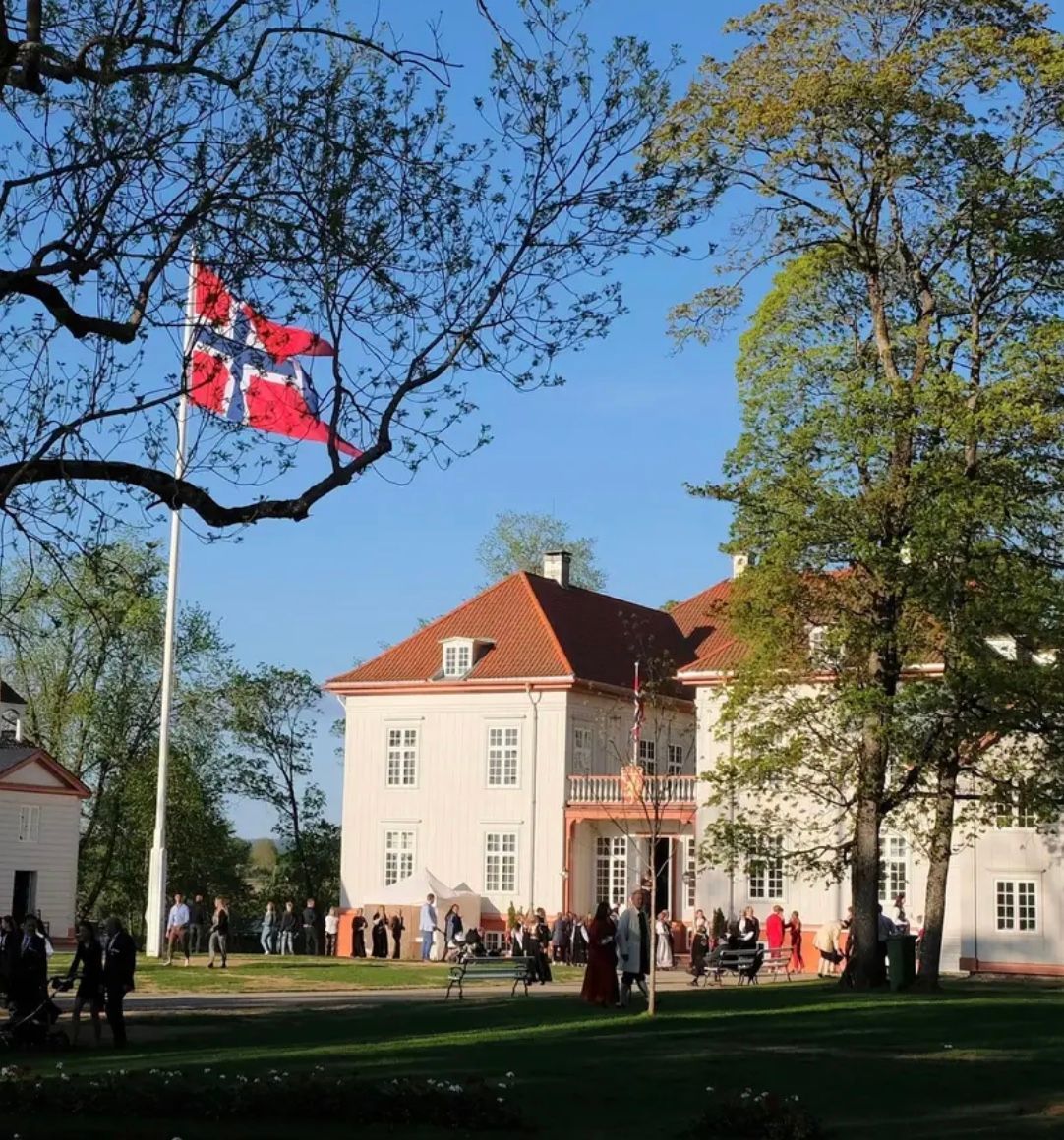 Et norsk flagg vaier over et hvitt herregårdshus i flere etasjer omgitt av trær på en solrik dag med folk samlet nedenfor.