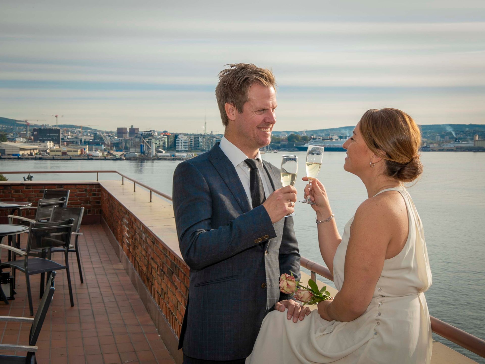 Et par i formelt antrekk skåler med champagneglass på balkong med utsikt over en byhavn ved solnedgang.