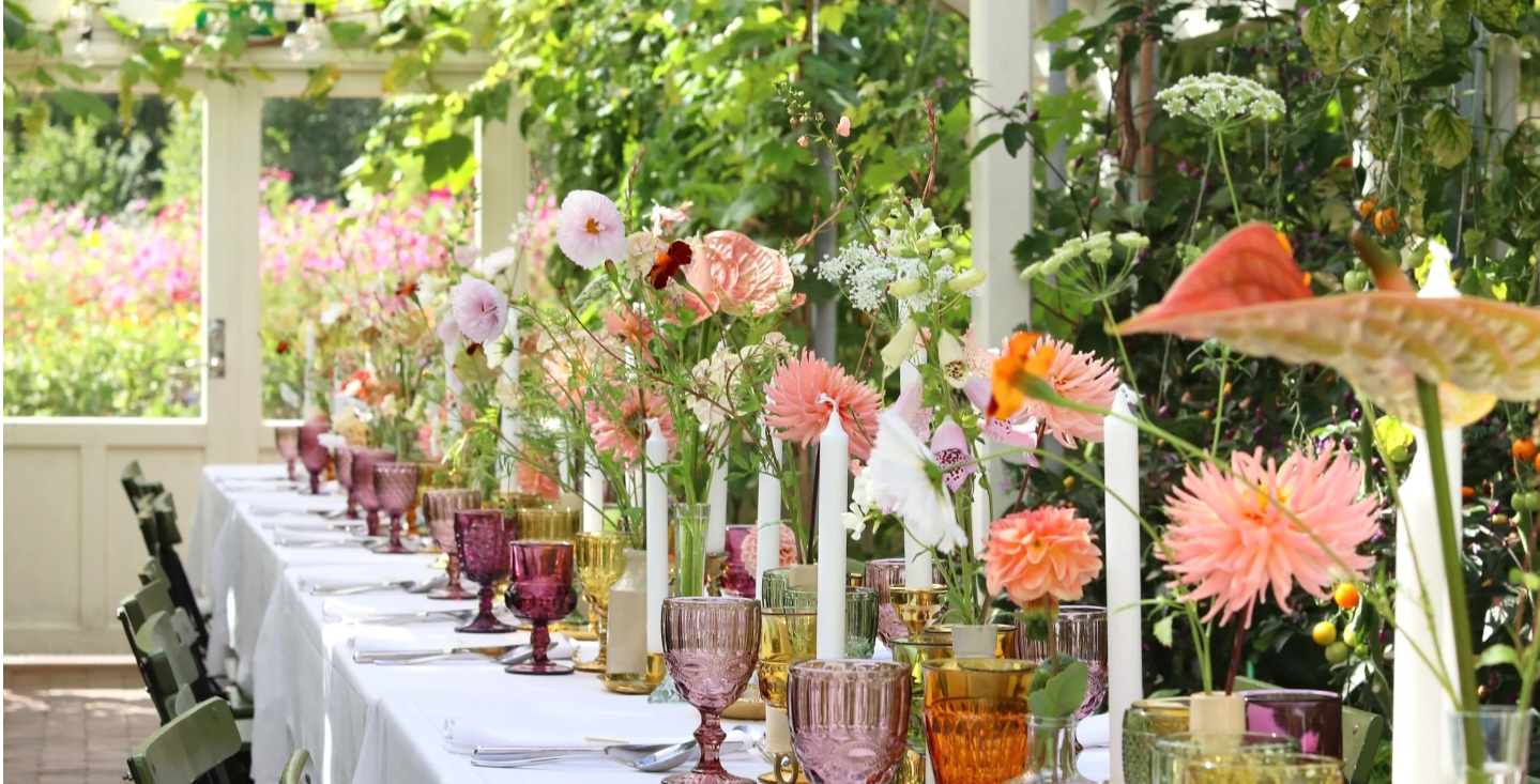 Et langt spisebord dekket med hvitt sengetøy, fargerikt glass, høye, slanke stearinlys og friske blomsterdekorasjoner.