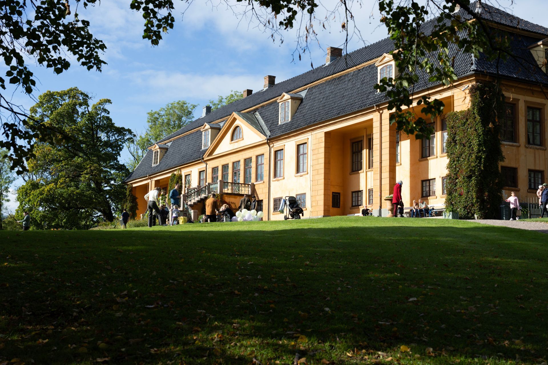 Et solrikt, langt gult herskapshus med mørkt tak og en gresskledd plen foran, sett fra en litt lav vinkel.