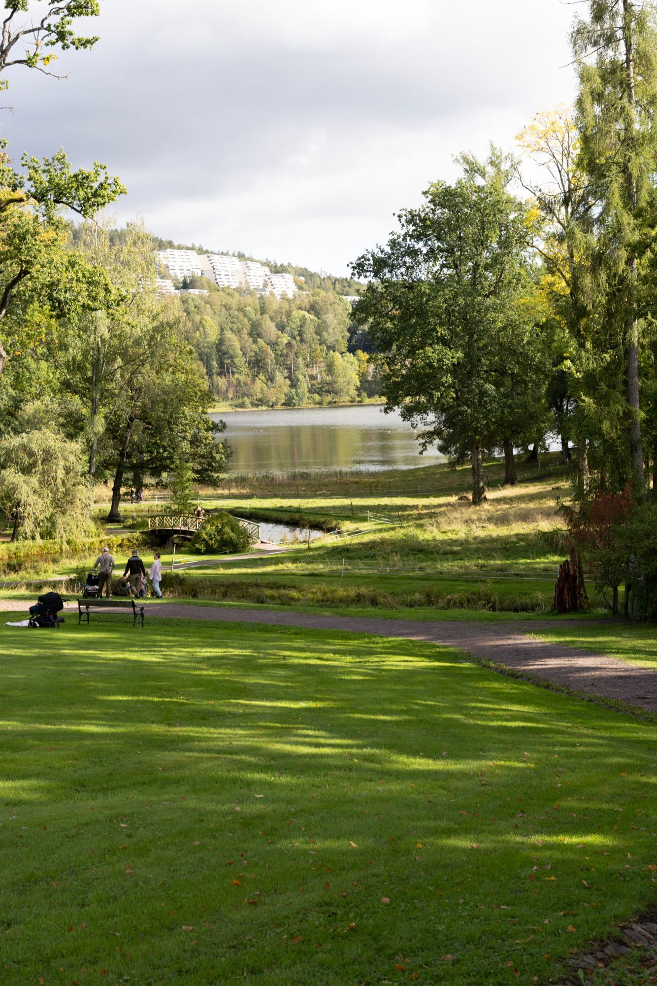 Parkplen og sti ved en innsjø, med folk som sitter under trær på en solskinnsdag