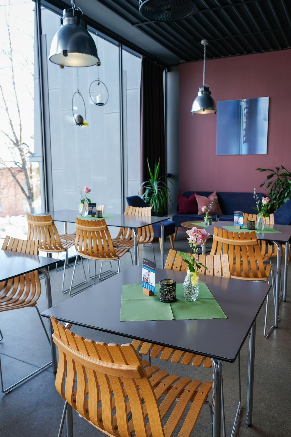 Moderne restaurantinteriør med lysebrune lamellstoler, grå bord, store vinduer og pendelbelysning.